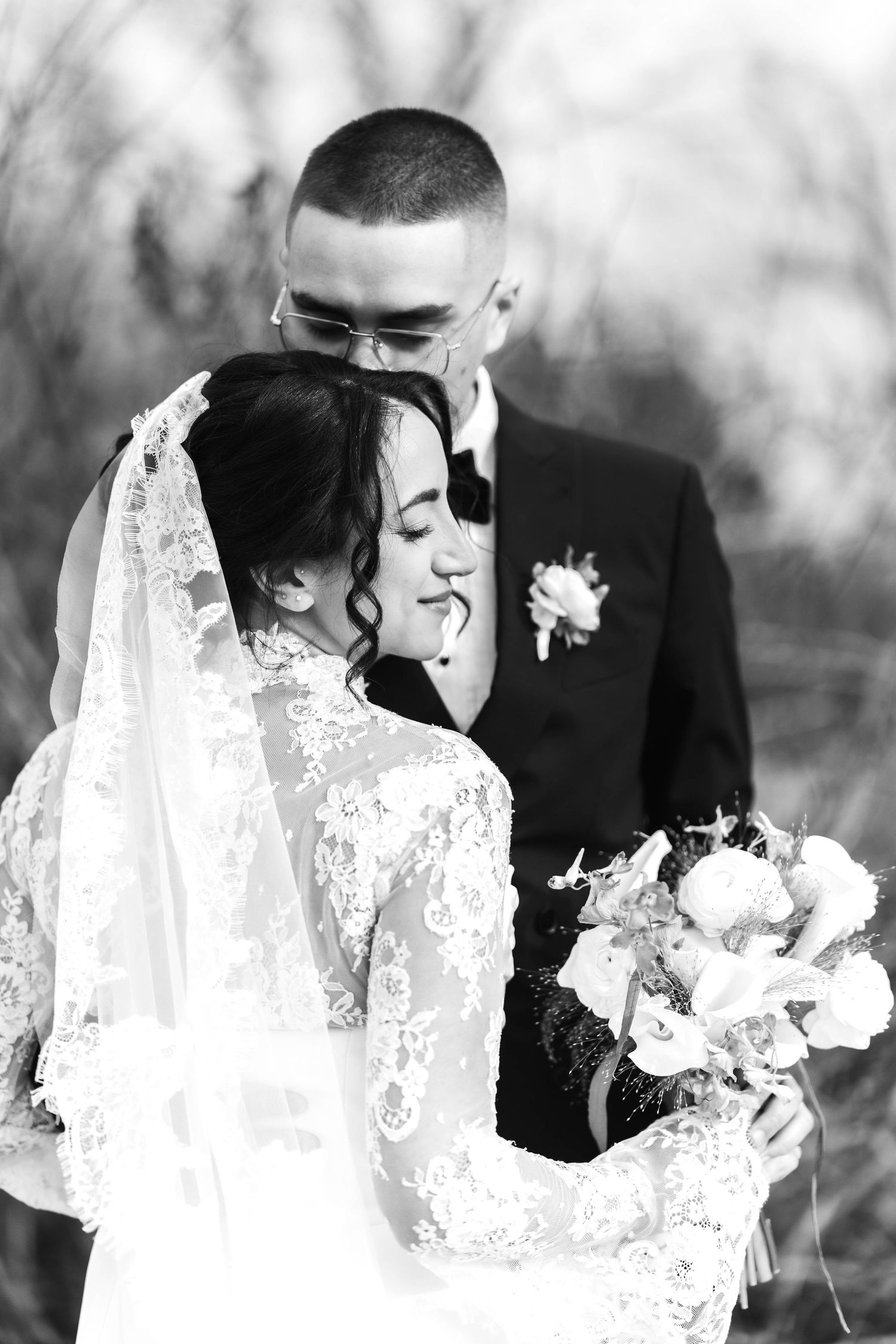 Black and white wedding portrait of bride in lace dress holding bouquet while groom in suit nuzzles her.