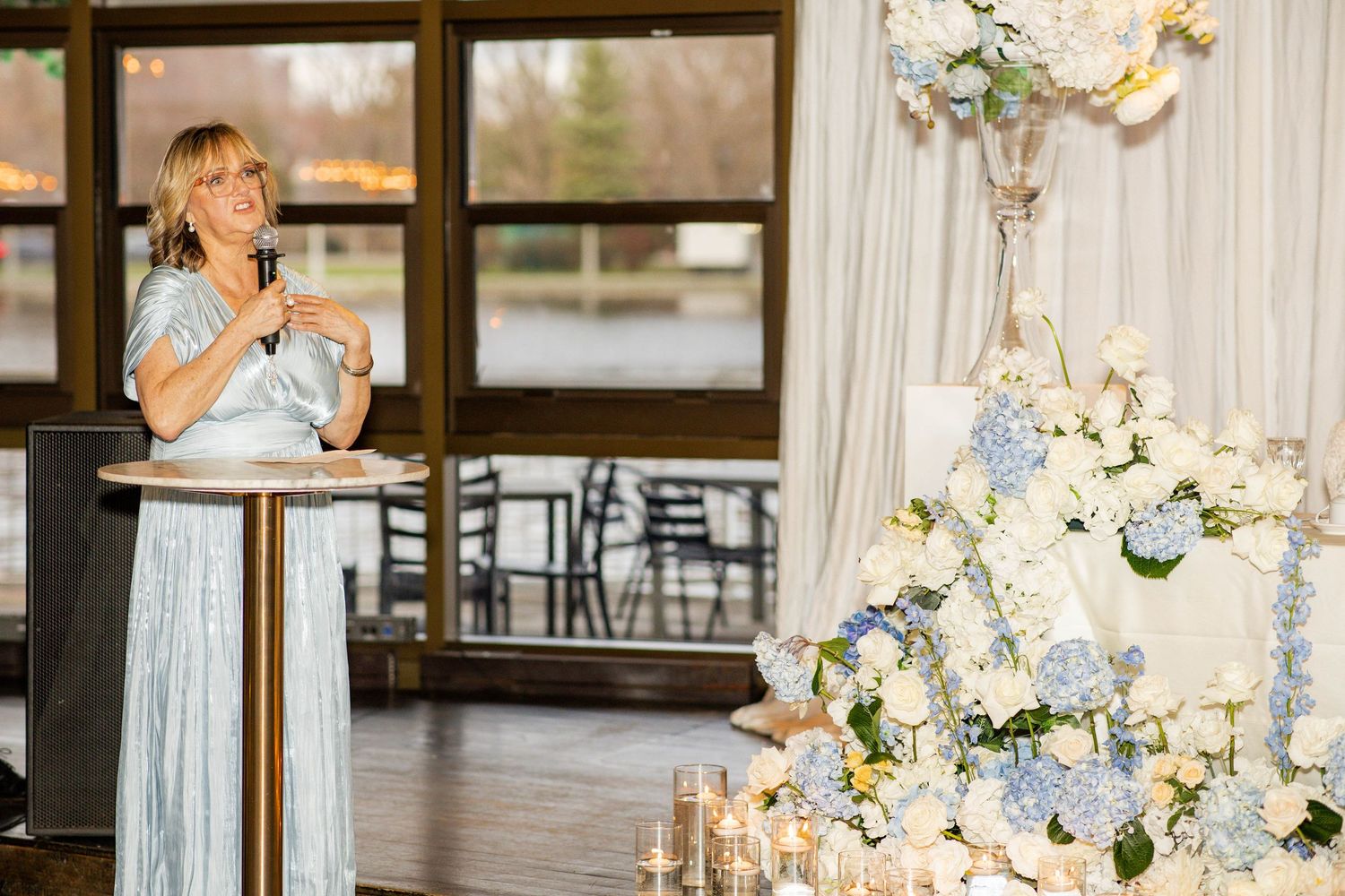 Woman in blue dress giving speech at elegant wedding reception with white floral cake and candles.