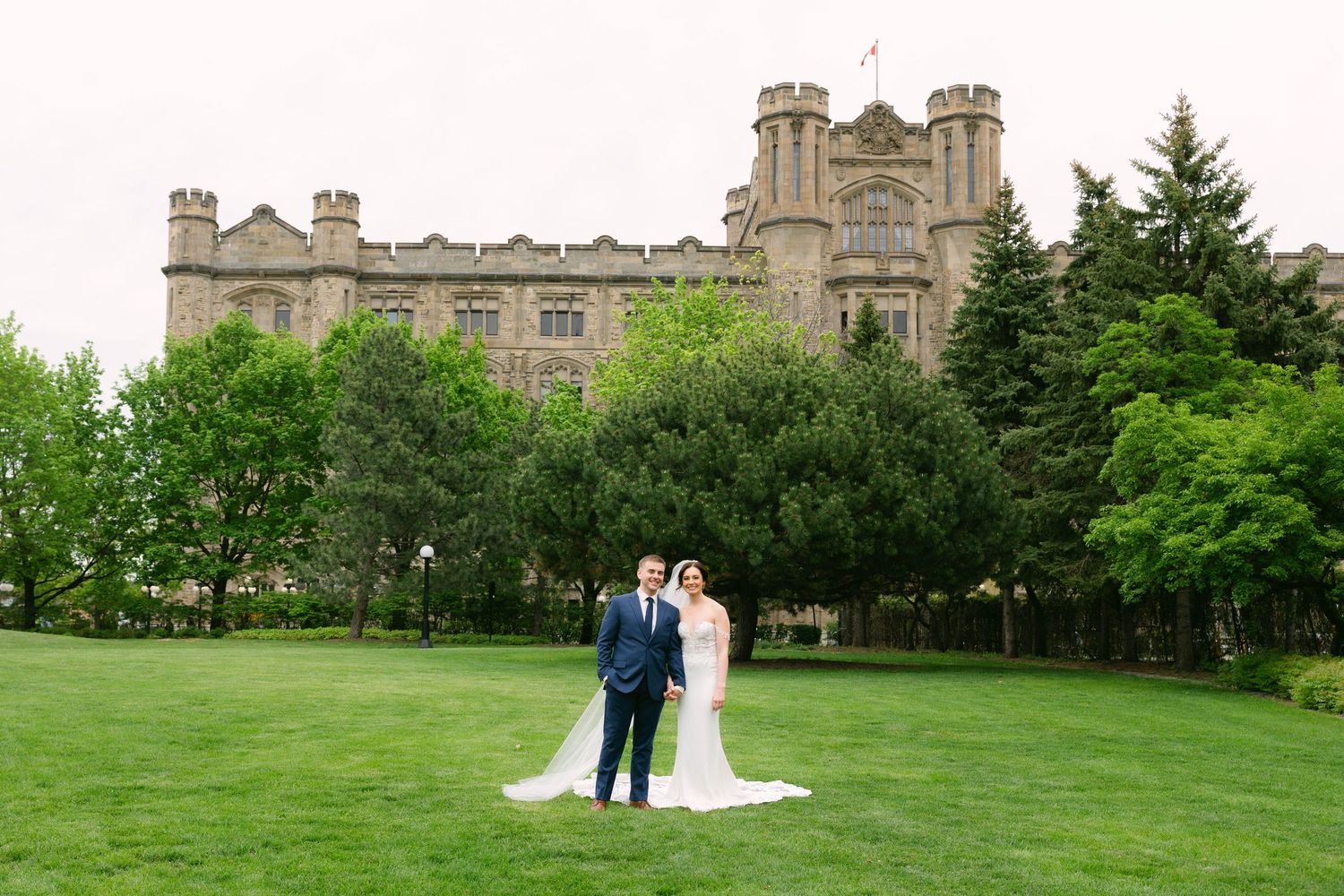 Bride and groom posing on lush green lawn before a grand castle-style building on their wedding day.