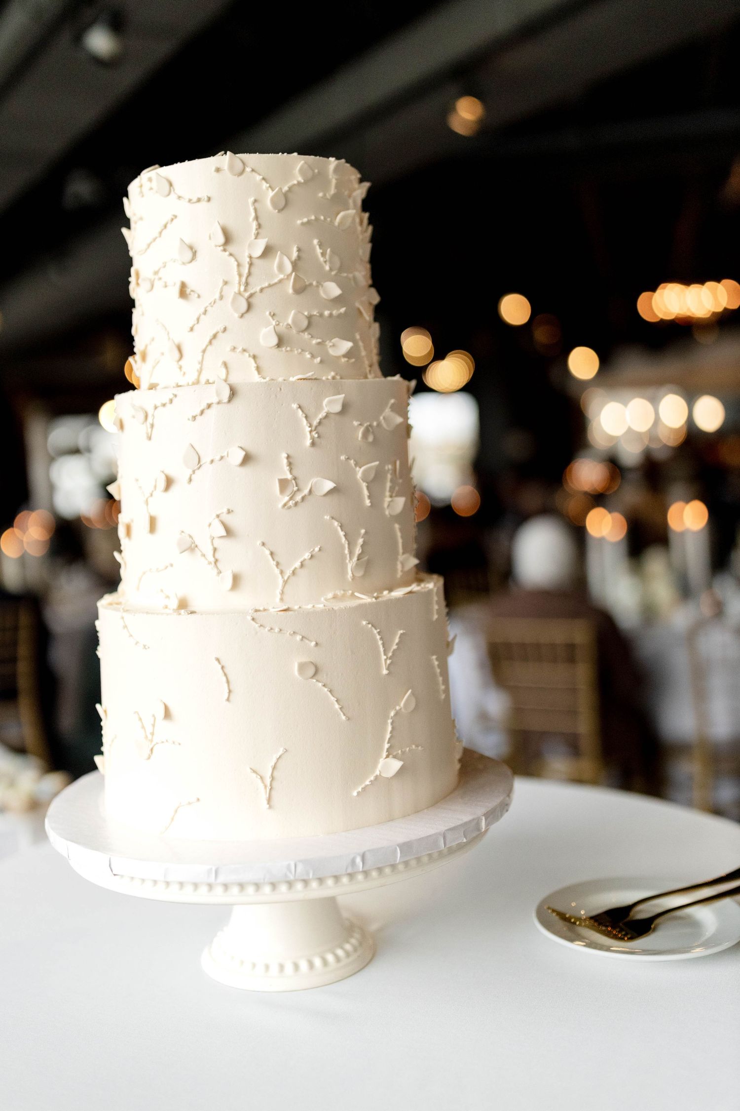 Three-tier white wedding cake with delicate floral buttercream details on a white pedestal stand with bokeh lights.