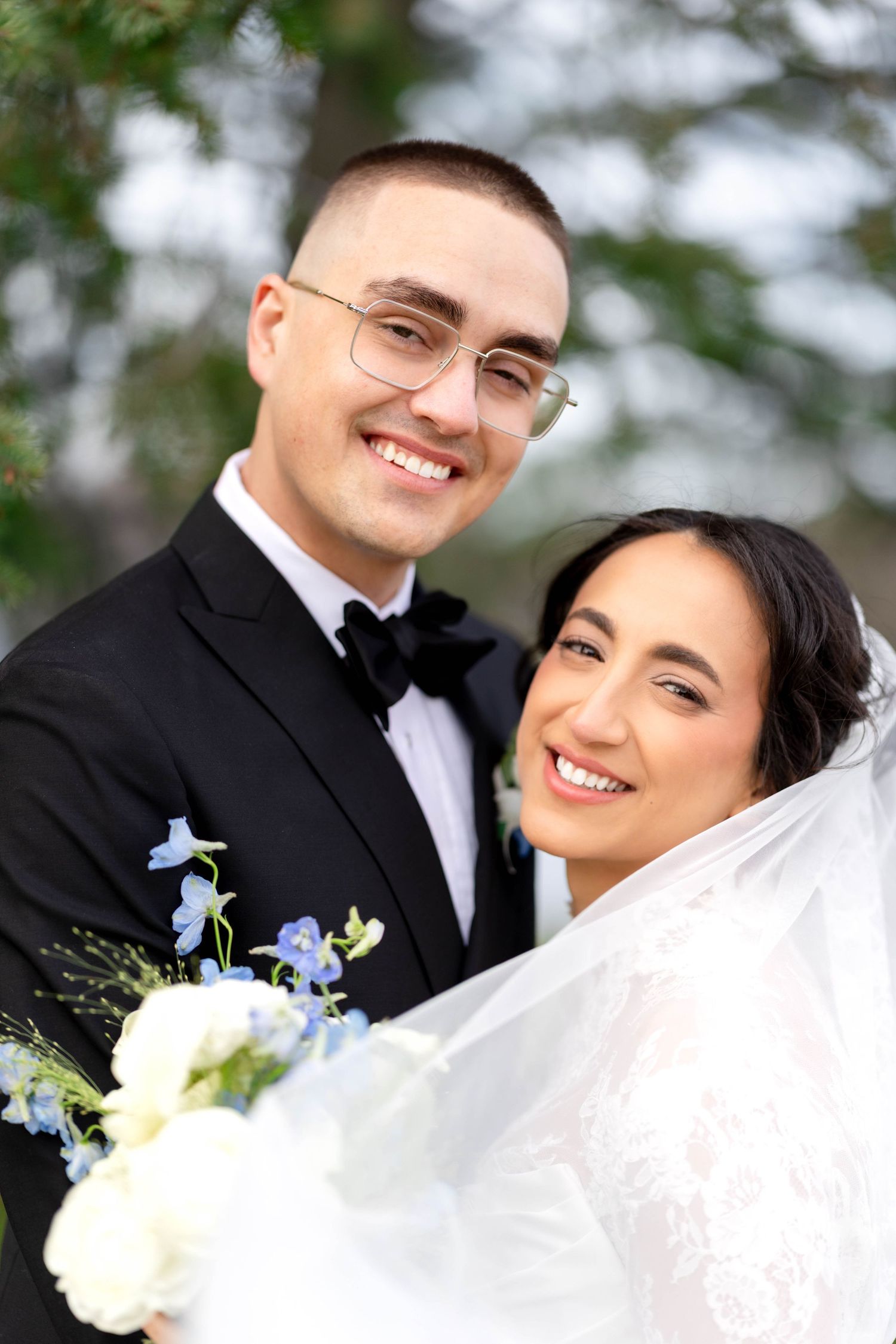 Smiling bride and groom pose outdoors, groom in black tuxedo with glasses, bride in lace dress holding floral bouquet.