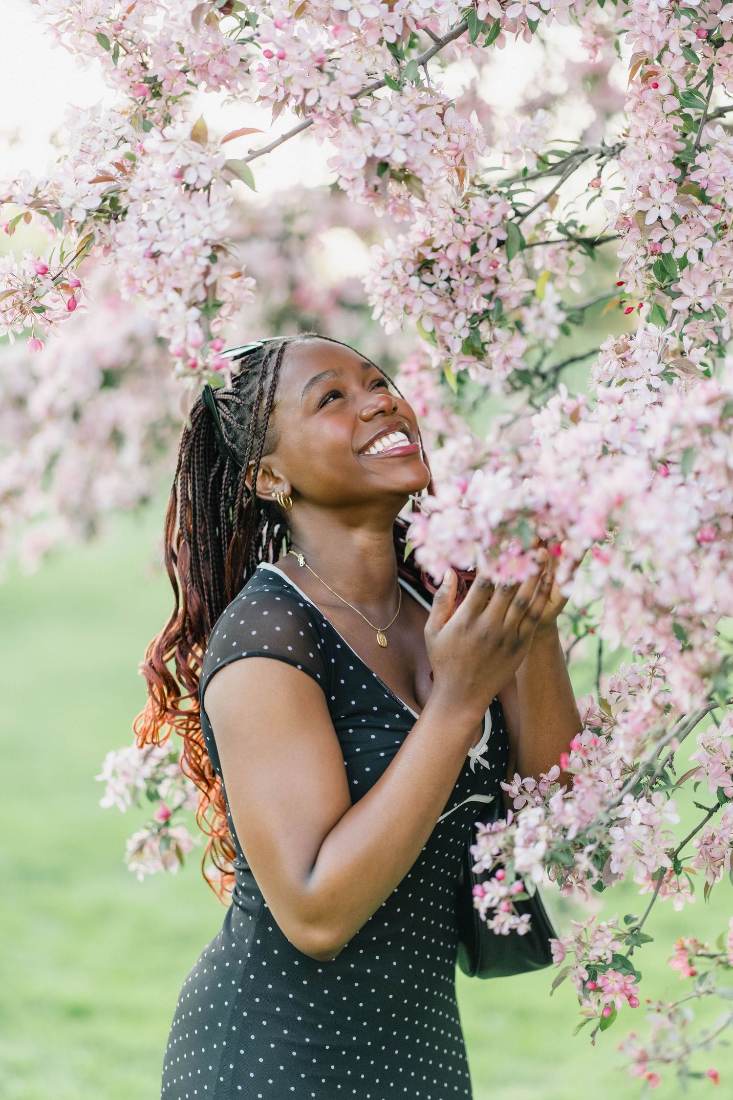 Smiling Black woman in polka dot dress touching pink cherry blossoms on tree branch in green park setting.