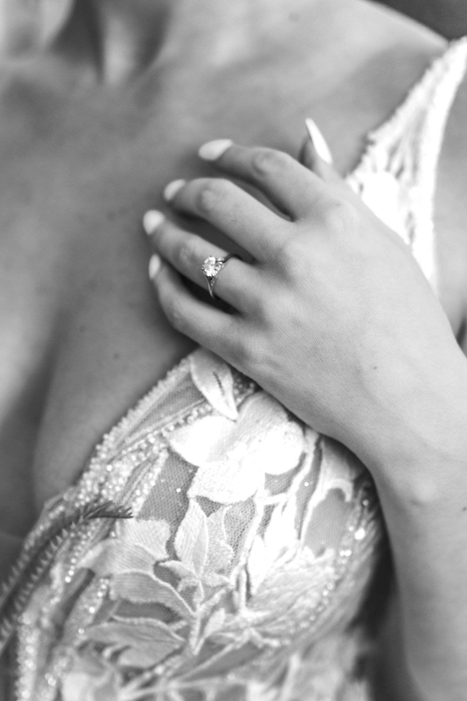 Black and white close-up image of hands gently touching an ornate lace wedding dress with delicate floral patterns.