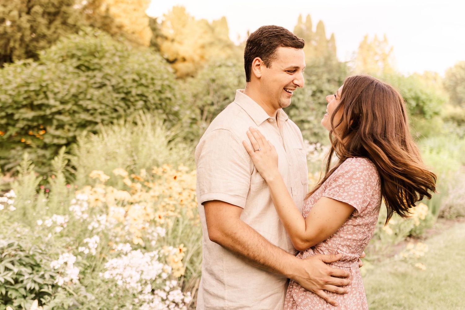 A couple embraces in a romantic outdoor setting during golden hour with wildflowers and natural scenery.