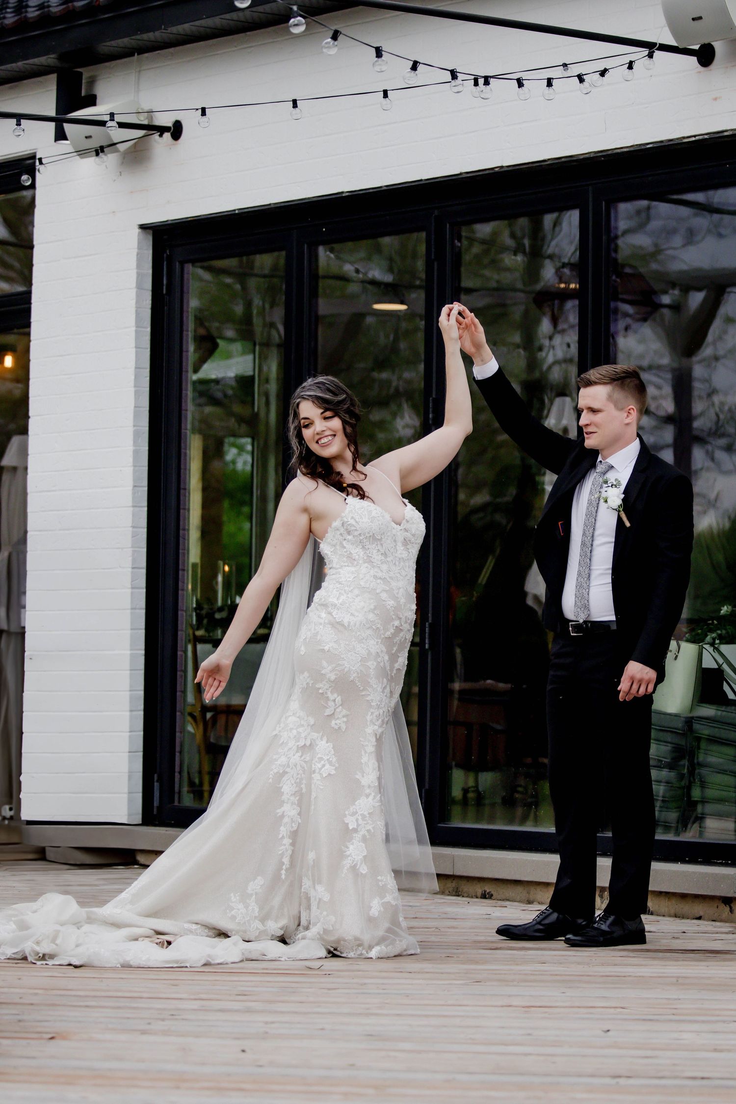 Newlyweds dance playfully on an outdoor deck with string lights and windows in the background.