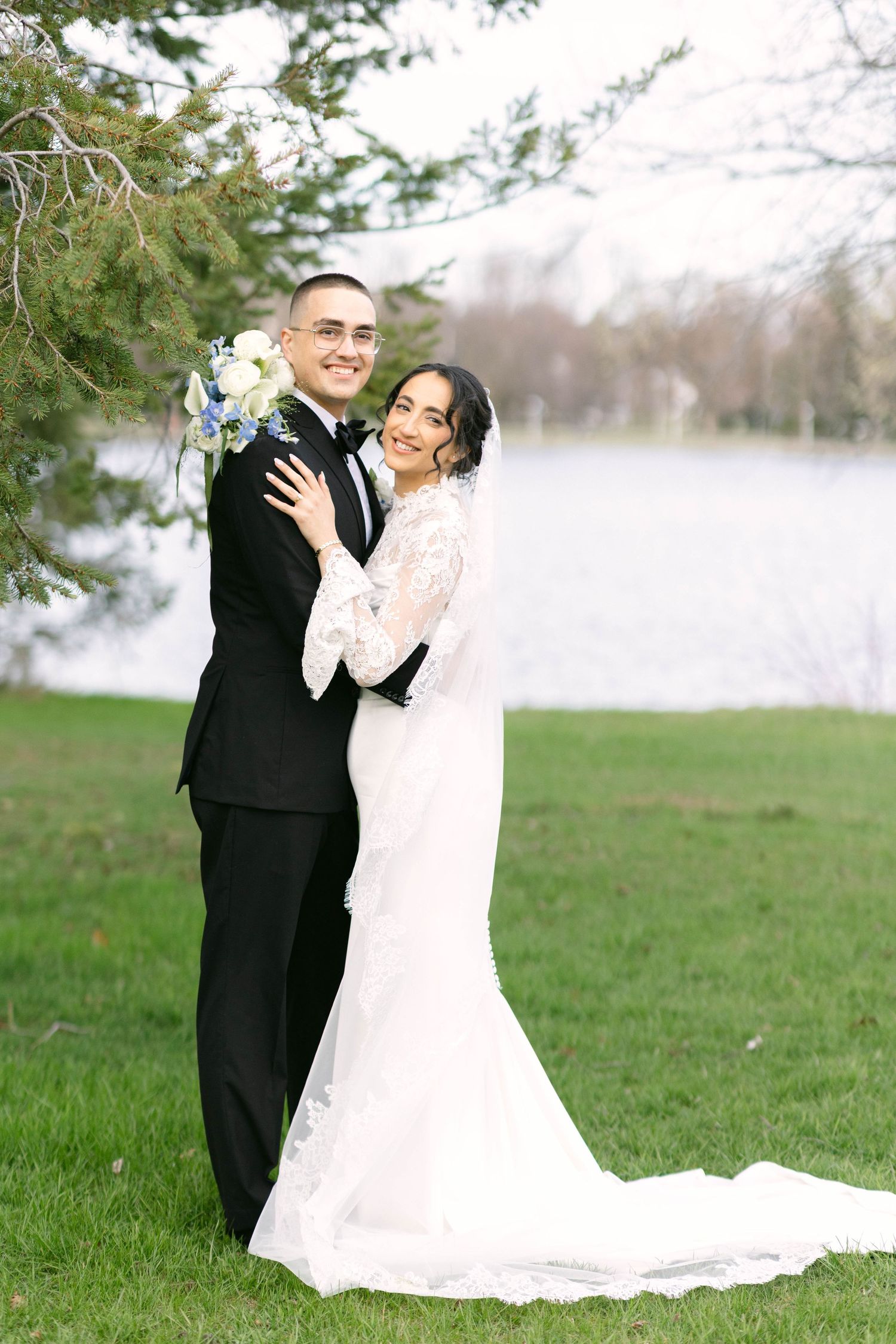 Bride in lace gown and groom in black tuxedo embrace near a lakeside tree on their wedding day.