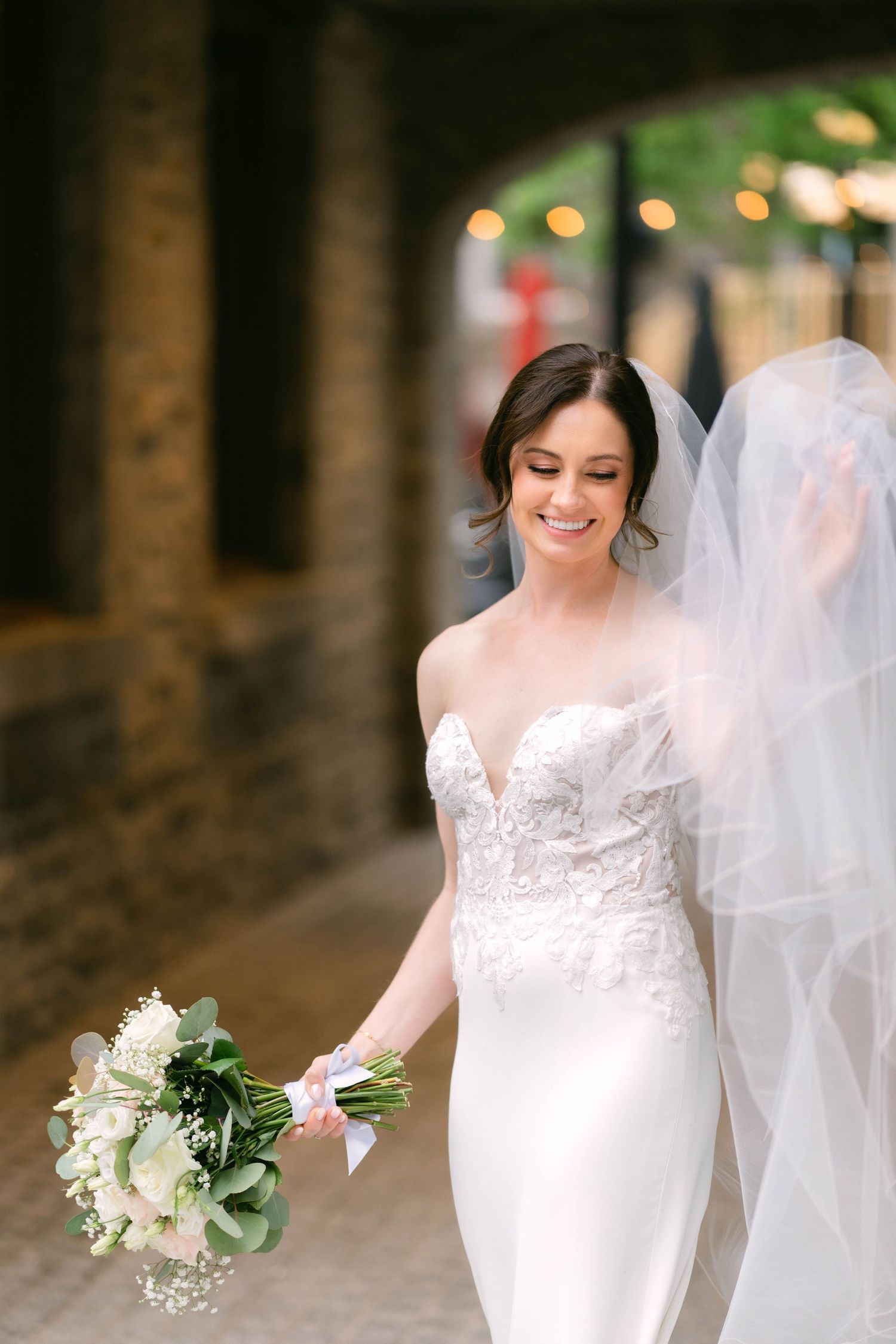 Smiling bride in lace strapless gown holds white bouquet, veil flowing, walking through elegant stone archway.