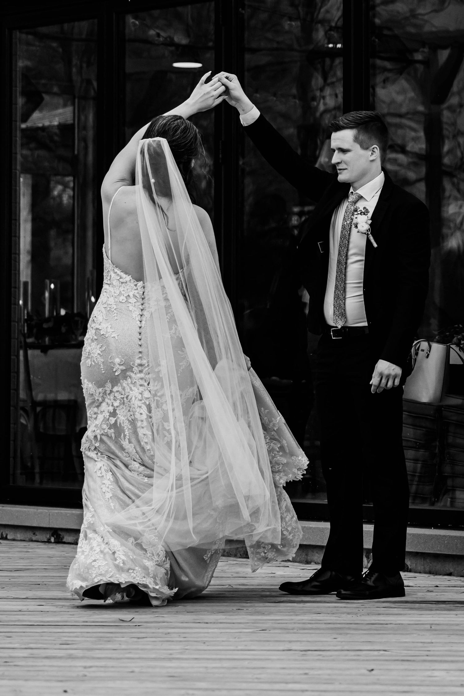 Black and white photo of newlyweds dancing together on a wooden deck outside a glass building.