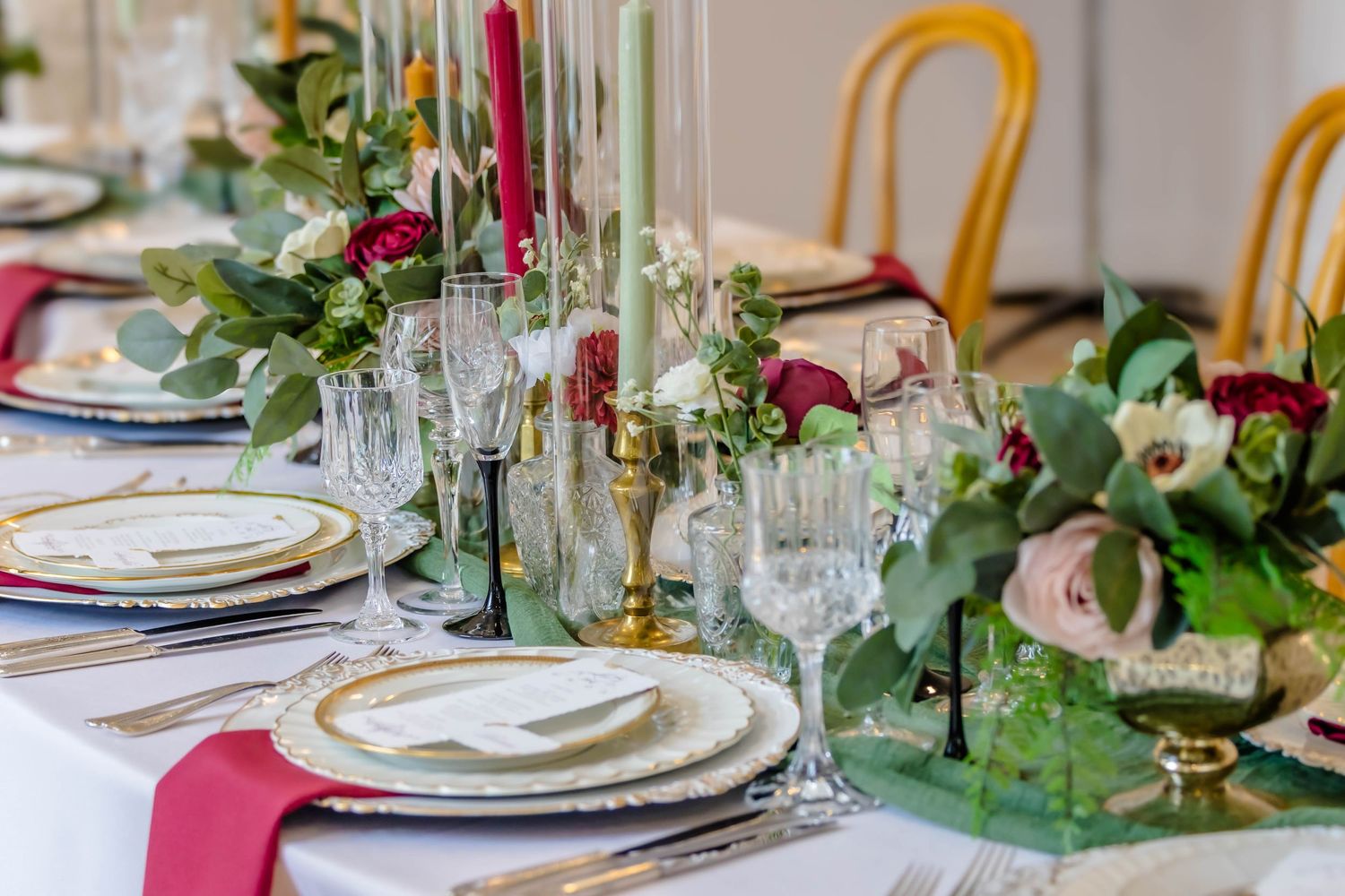 Formal wedding table setting with crystal glasses, gold rimmed plates, and burgundy floral centerpieces.