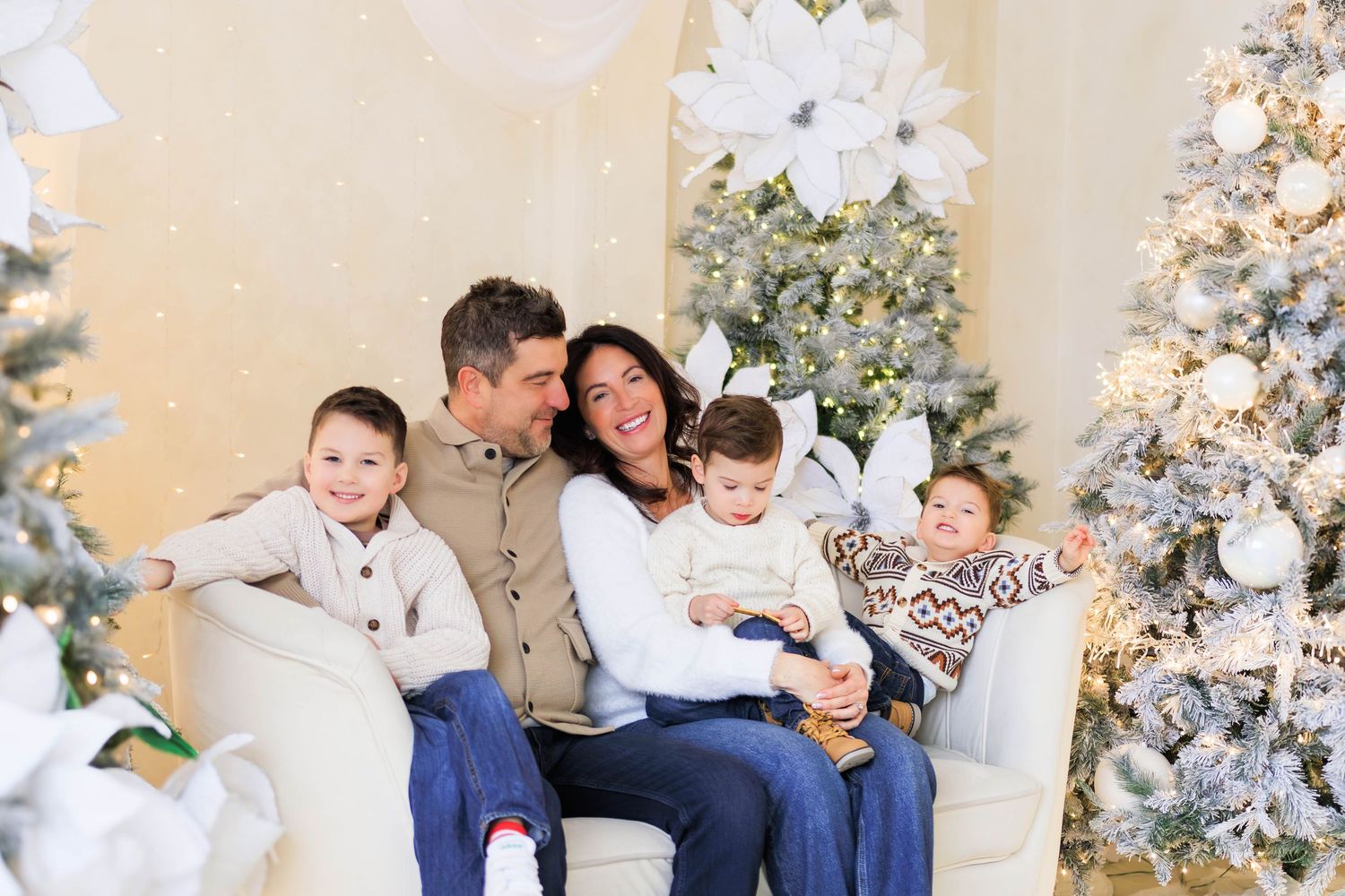 A joyful family sitting together on a white couch surrounded by decorated Christmas trees.
