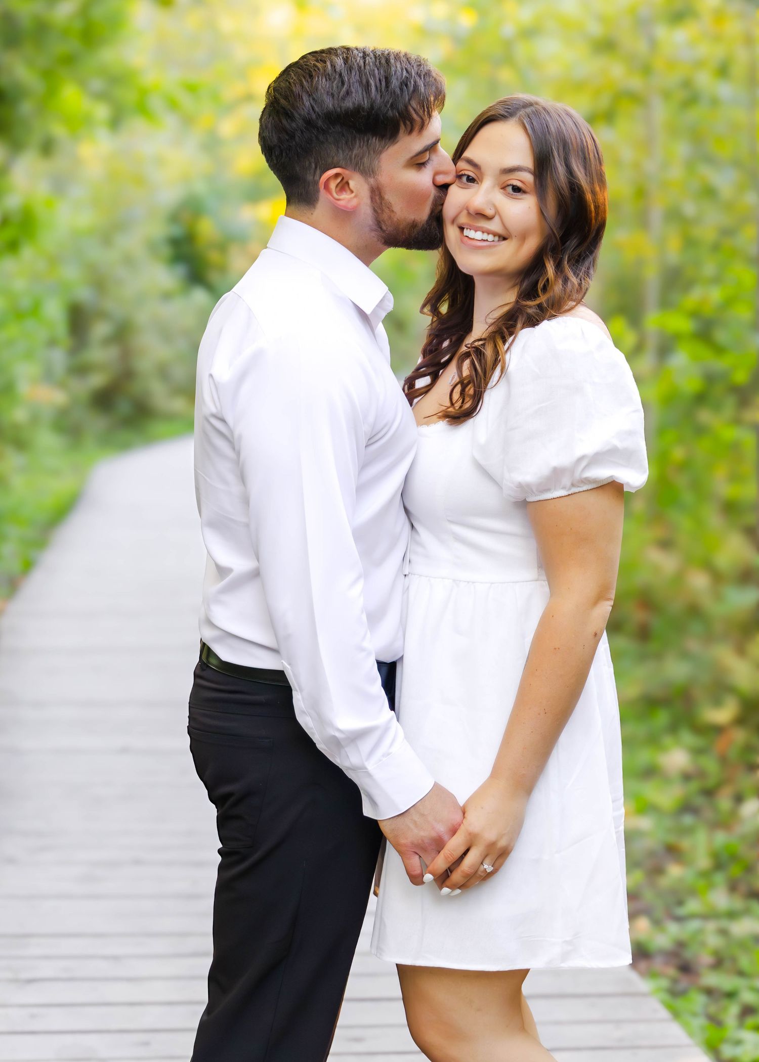 Intimate couple's photoshoot on wooden path with natural green backdrop during golden hour.