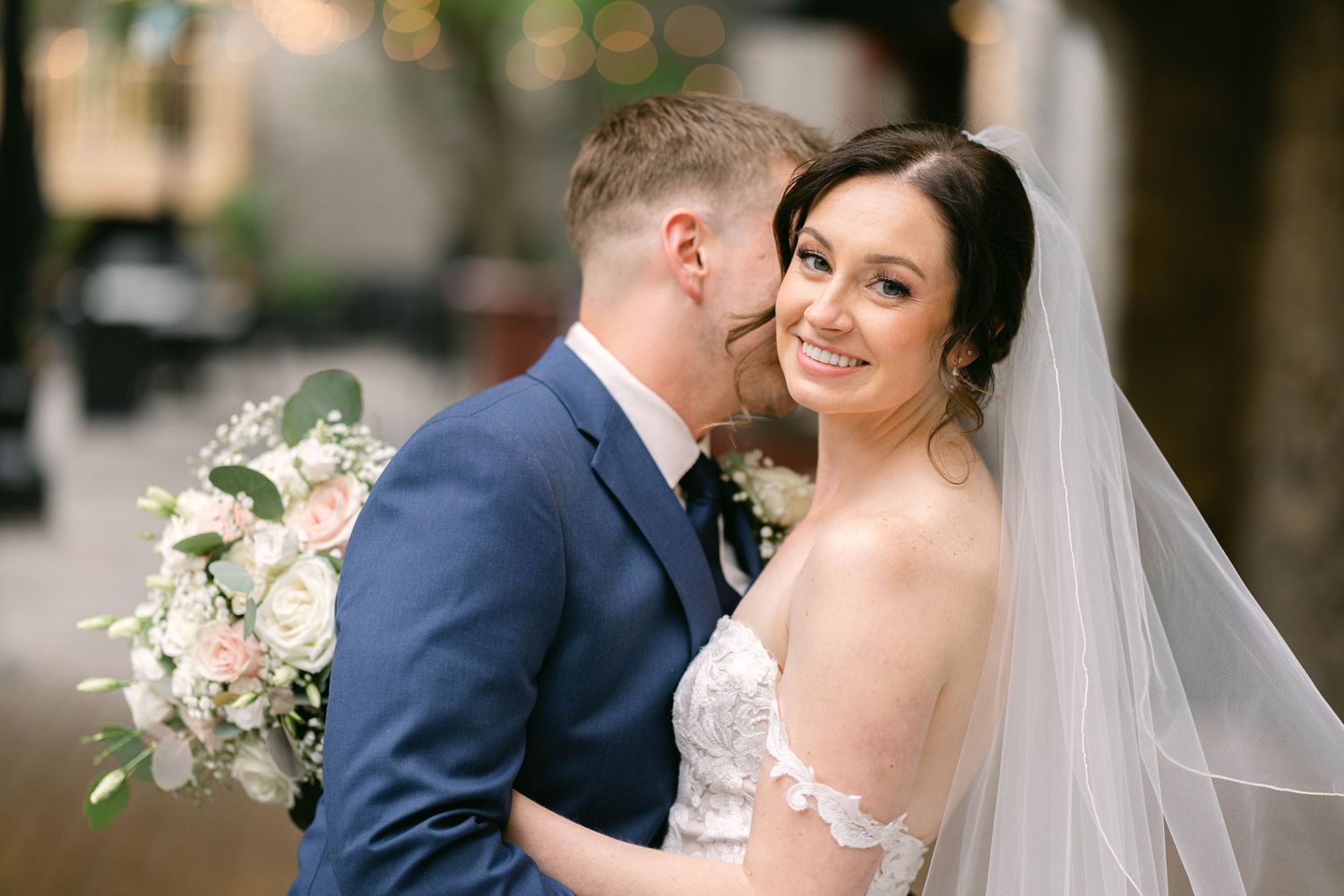 Bride in lace gown holding white floral bouquet smiles as groom in navy suit embraces her at wedding.