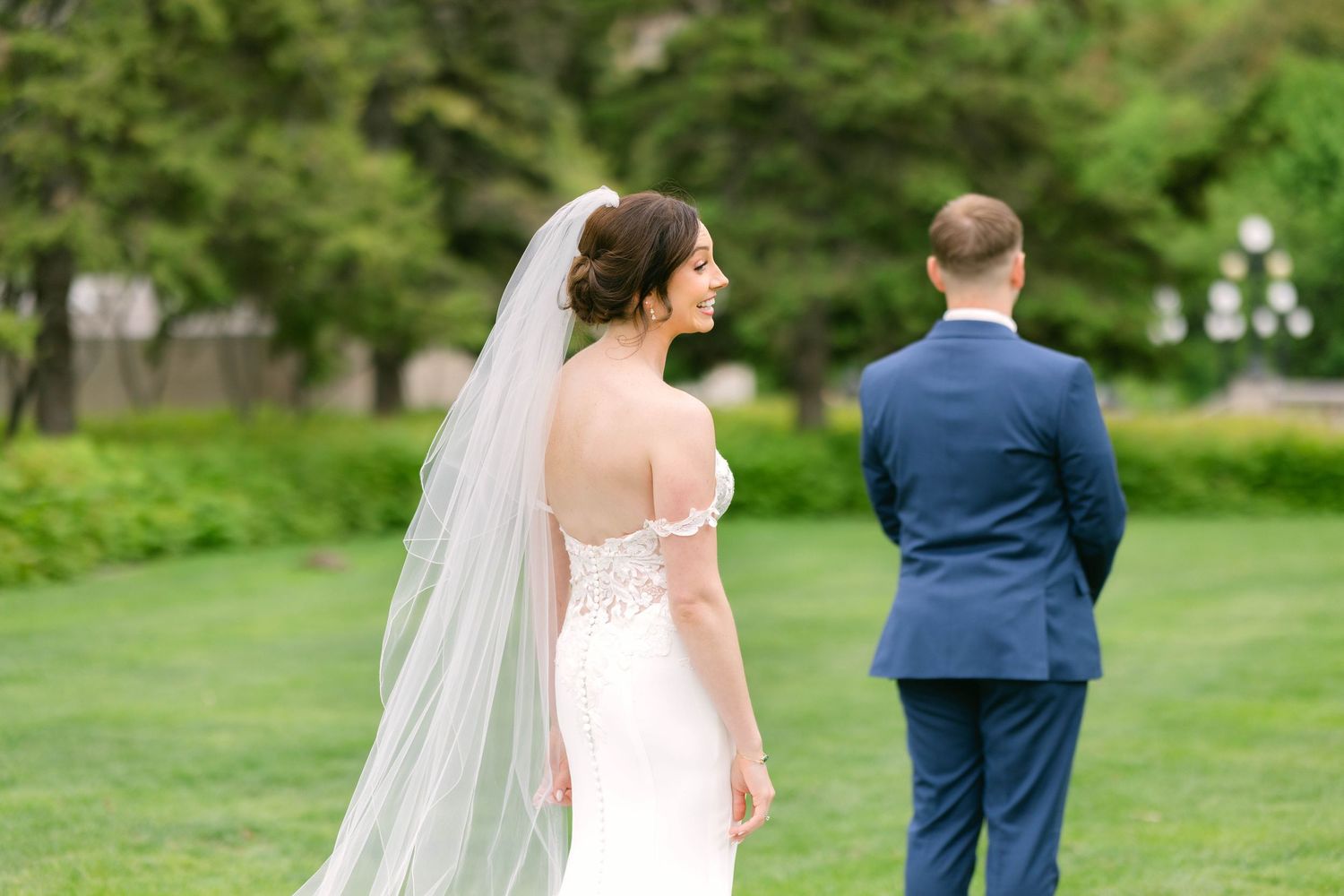 Bride in lace wedding gown and veil stands behind groom in navy suit during outdoor first look on green lawn.
