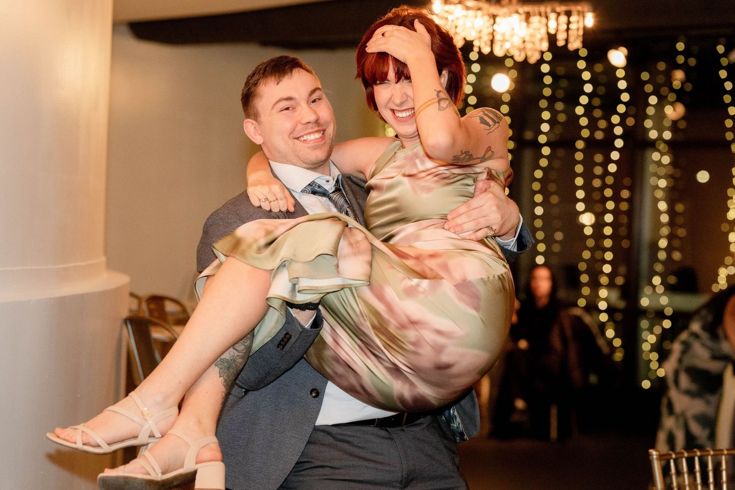 Groom carrying bride in elegant dress at wedding reception with bokeh string lights background.