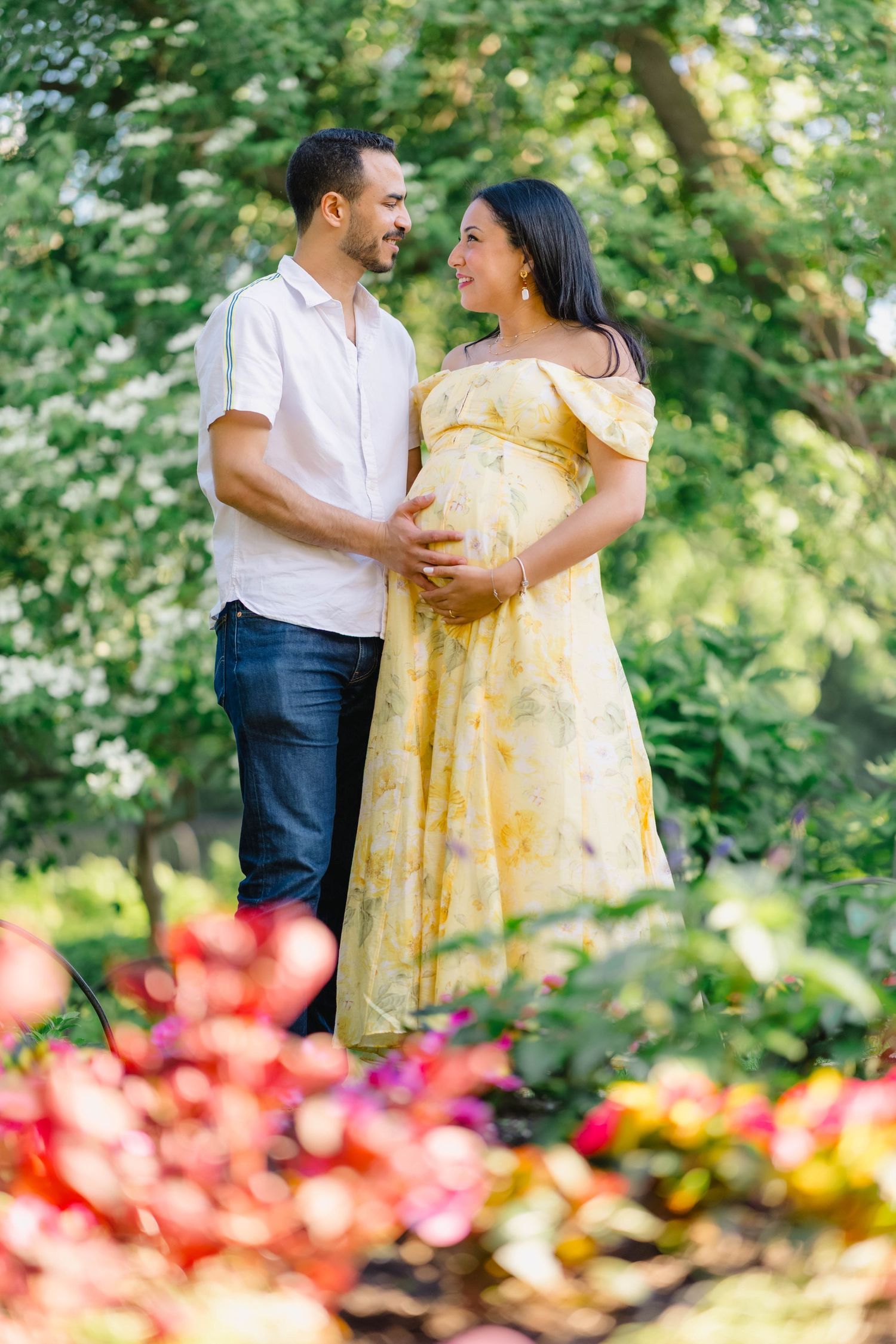 A couple shares a tender moment in a lush garden, the woman in a yellow floral maternity dress cradling her baby bump.