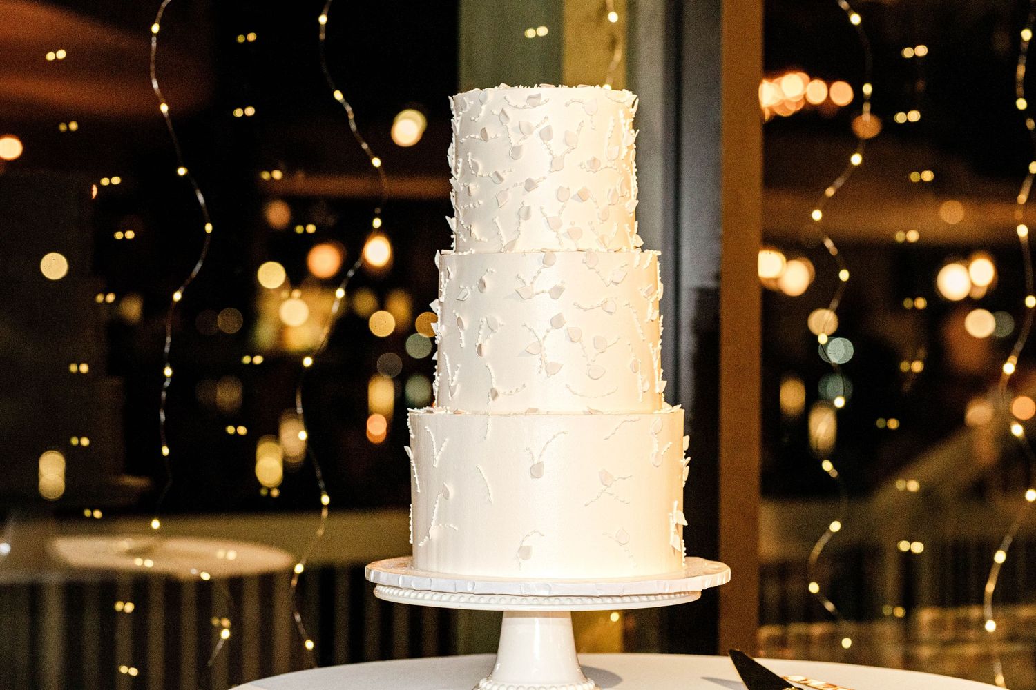Three-tier white wedding cake with textured frosting details on a white stand with bokeh lights in background.