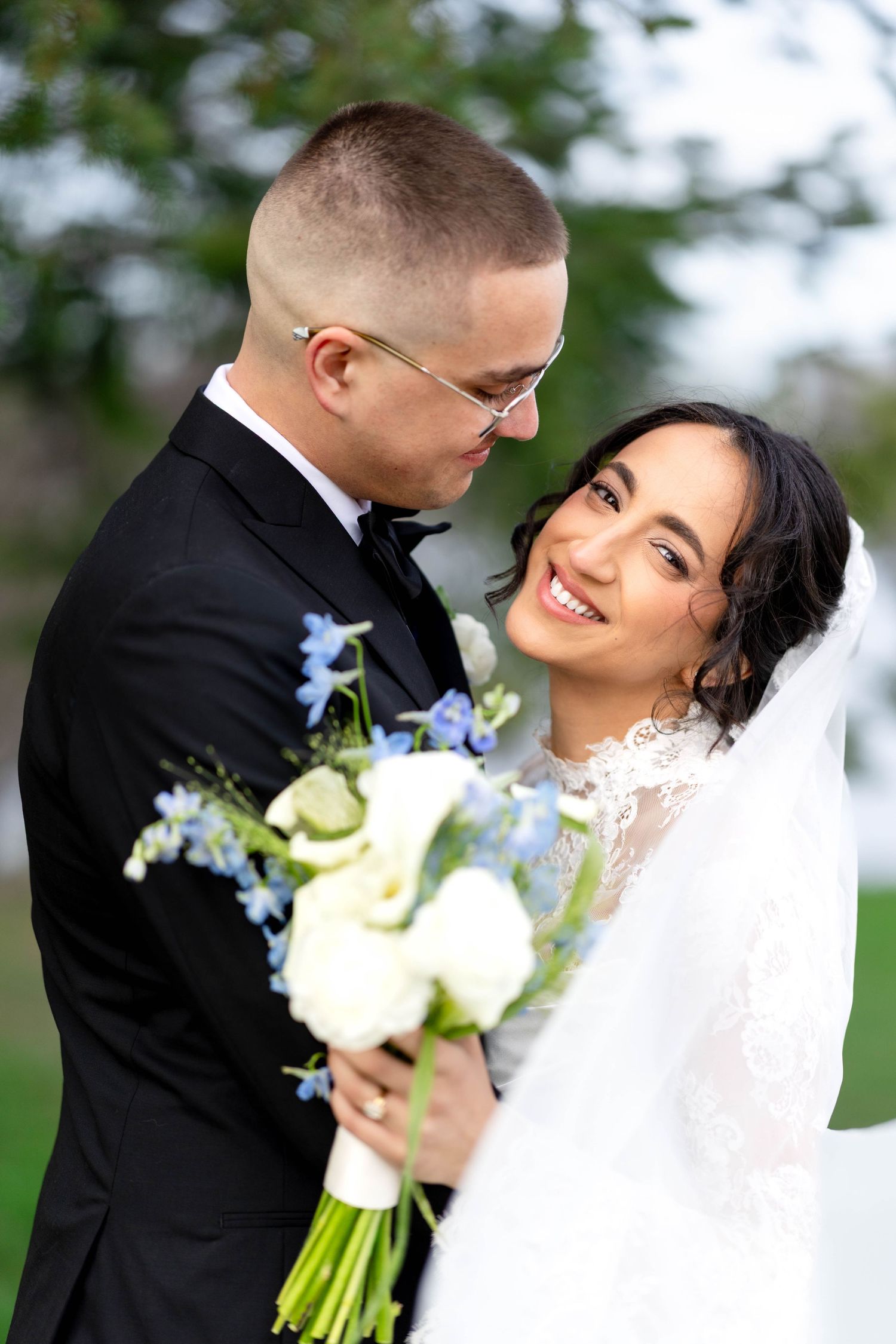 Smiling bride in white lace gown holds blue and white bouquet beside groom in black tuxedo outdoors.