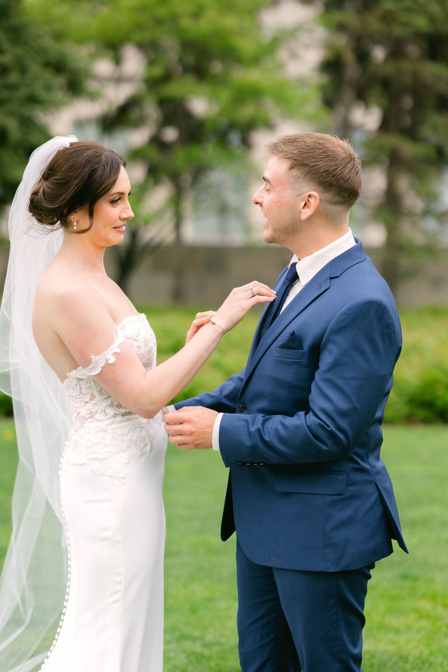 Bride and groom share a tender moment on green lawn, groom holding bride's hand in lace off-shoulder gown.