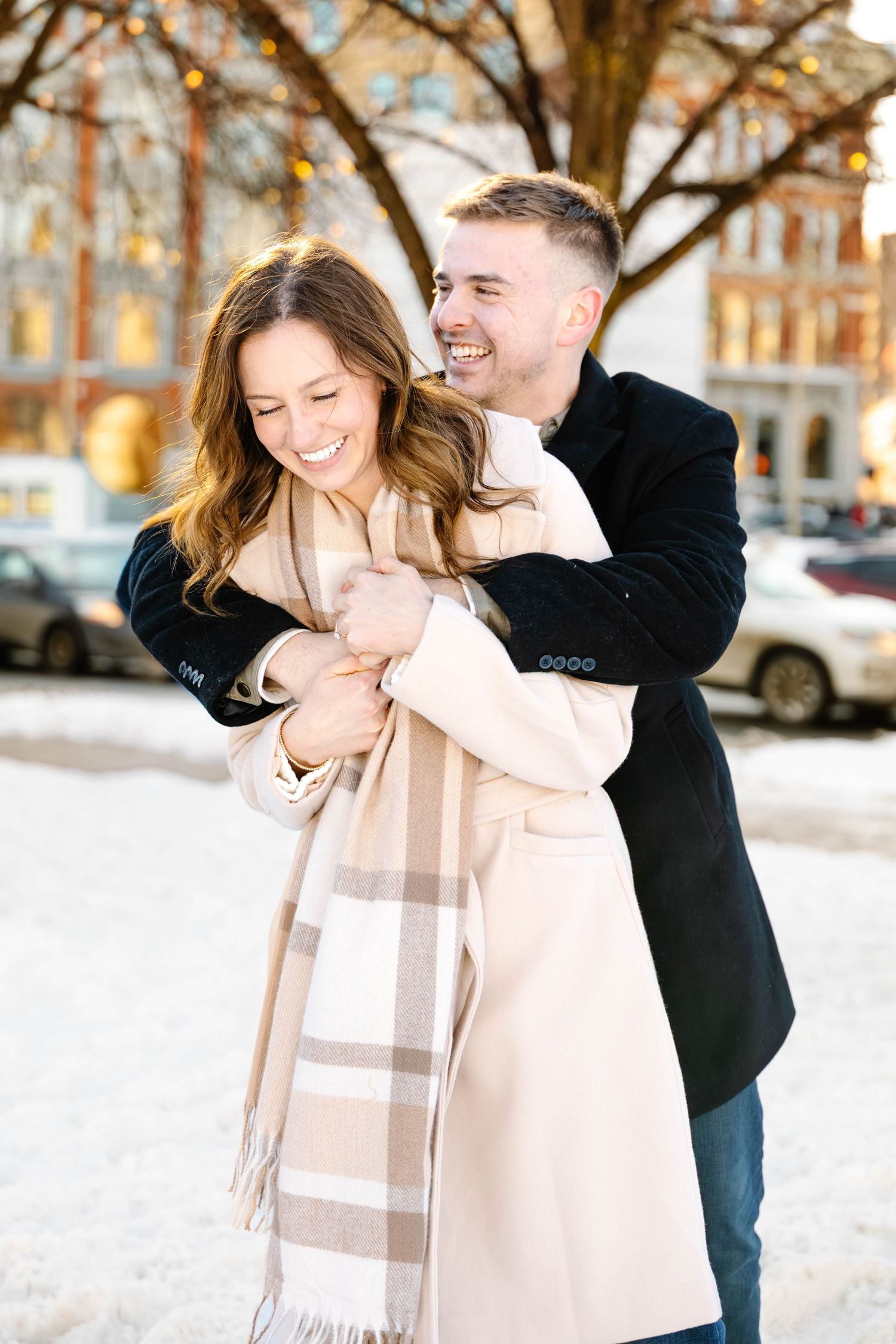 A couple embraces and laughs together in a snowy winter scene wearing a black coat and beige scarf.