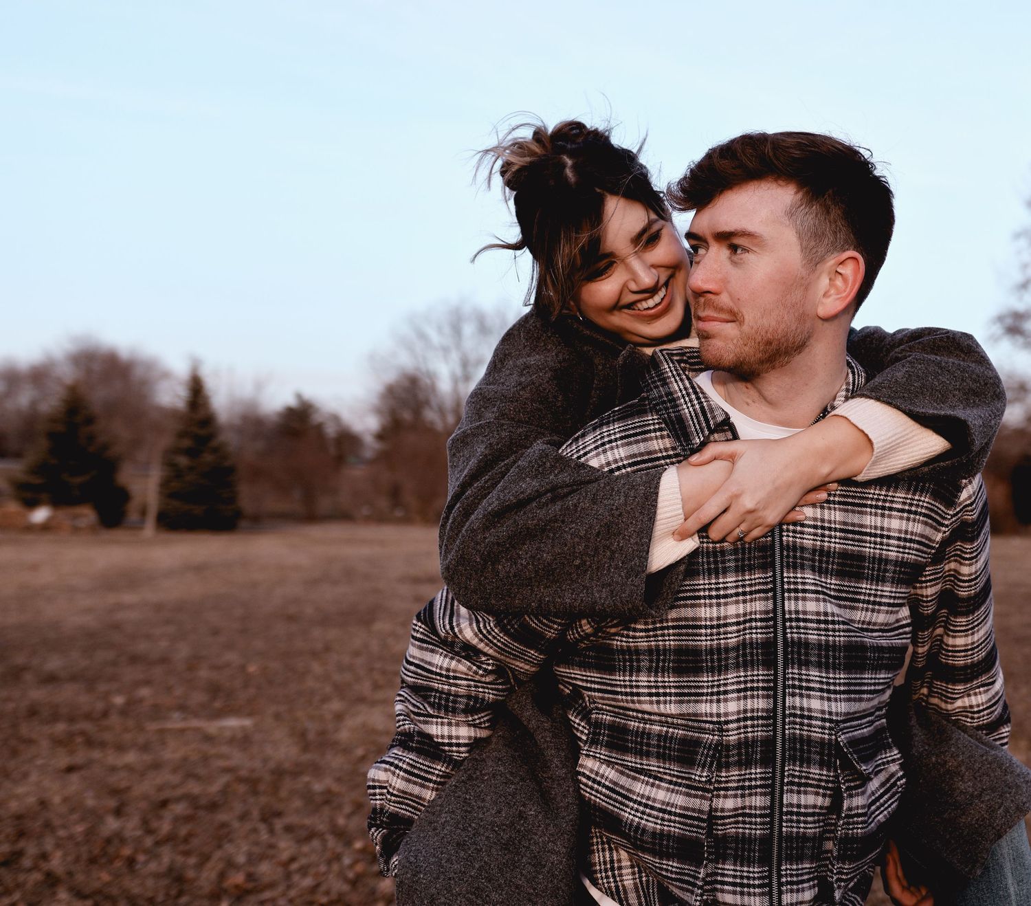 A couple wearing matching plaid shirts embrace lovingly in a rustic outdoor setting during golden hour.