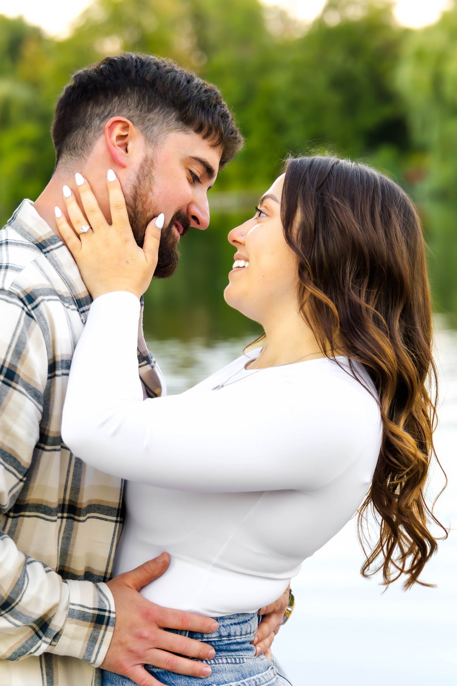 A couple laughs and embraces by a lake during a romantic outdoor photo session in natural lighting.