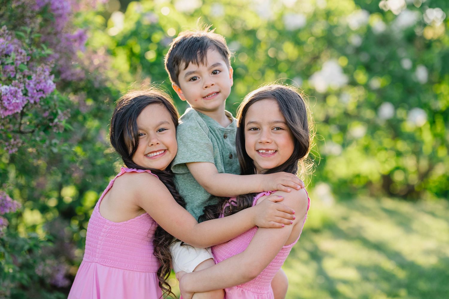 Three happy children hugging outdoors in a garden with blooming pink flowers and lush green trees.