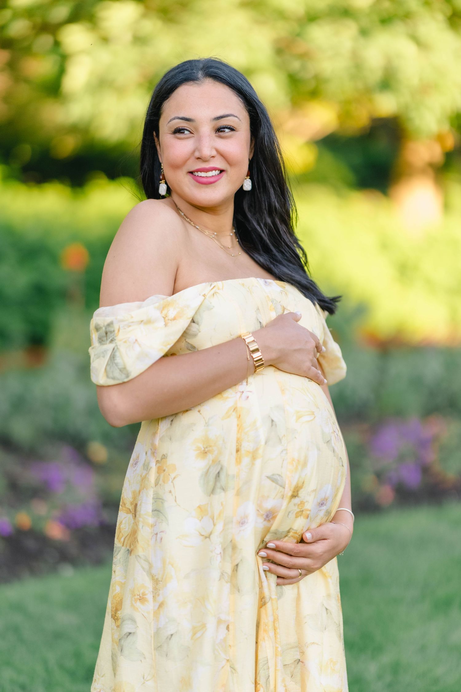 Smiling pregnant woman in yellow floral off-shoulder dress cradling baby bump in a lush garden setting.