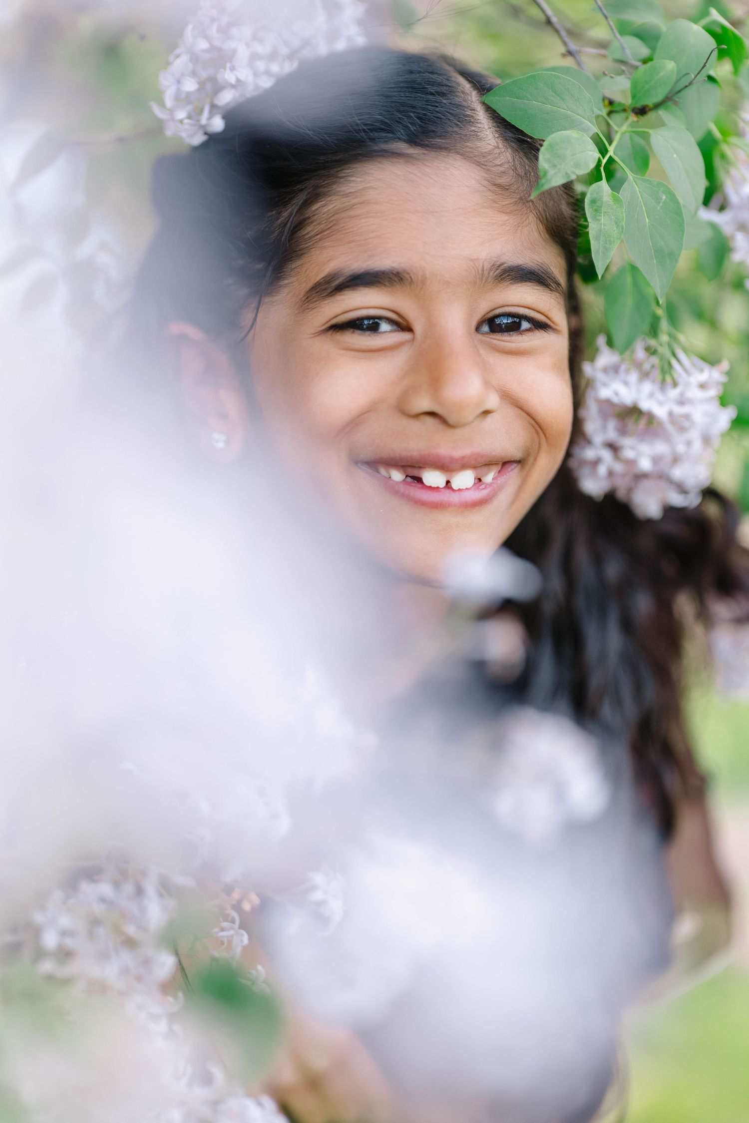 A smiling young girl peeks through blooming white lilac flowers and green leaves in a soft-focus outdoor portrait.