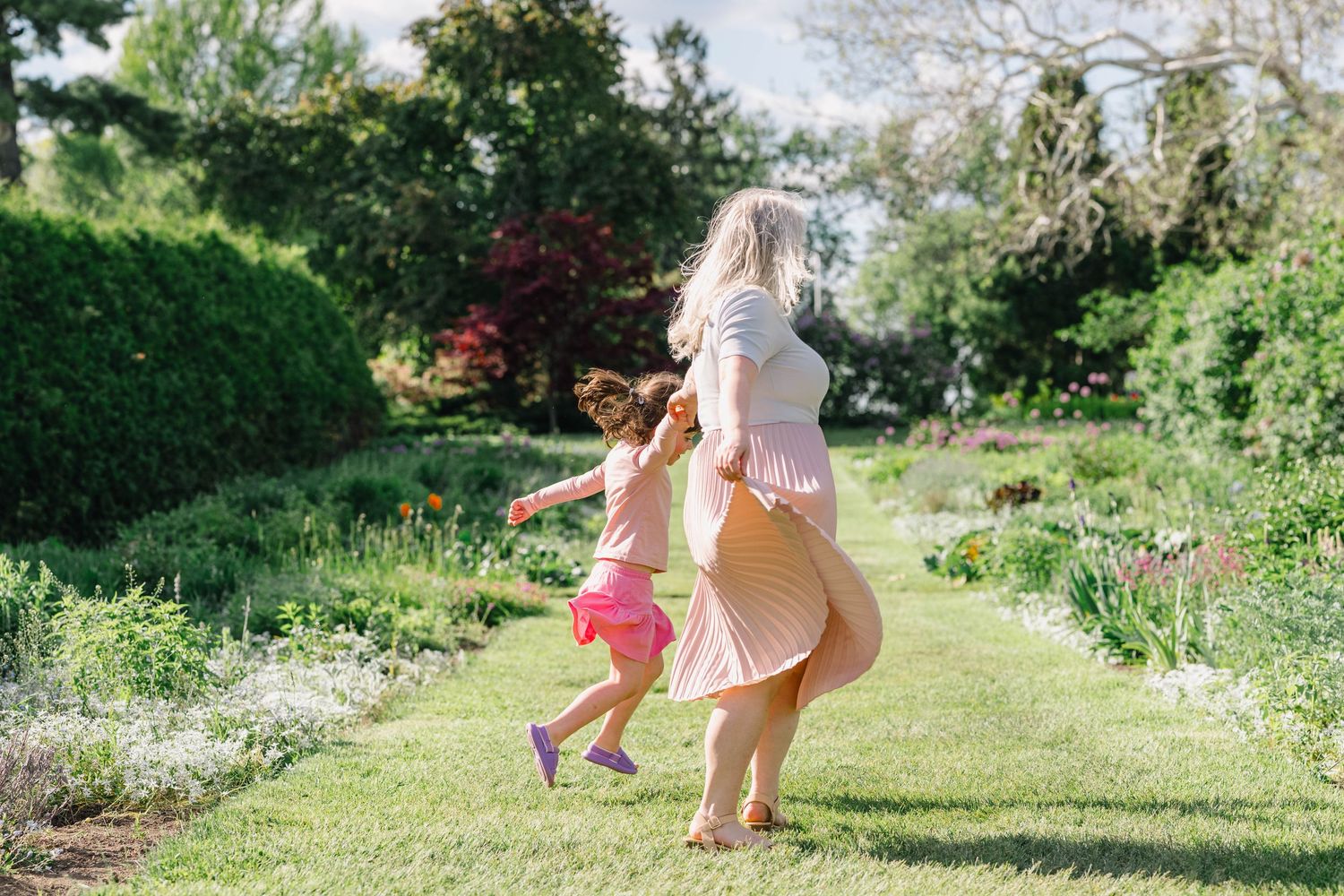 A woman and young girl run together through a lush green garden path on a sunny day.