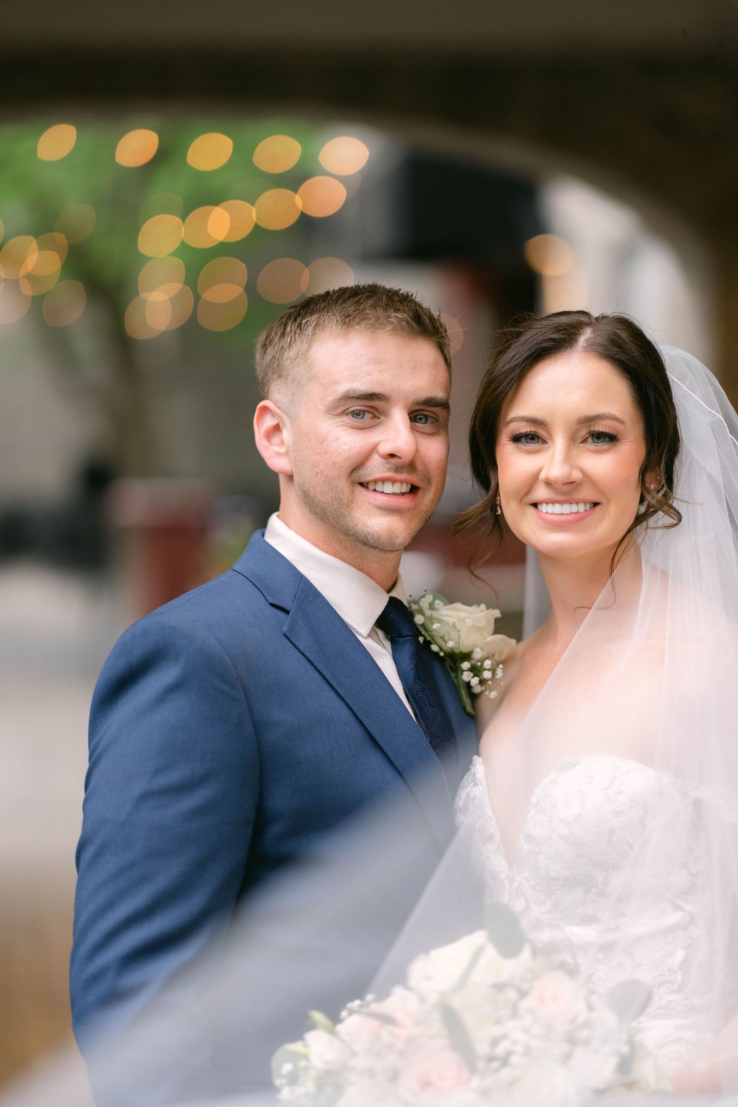 Smiling bride and groom pose together, she in a lace gown with veil, he in a navy suit with boutonniere.