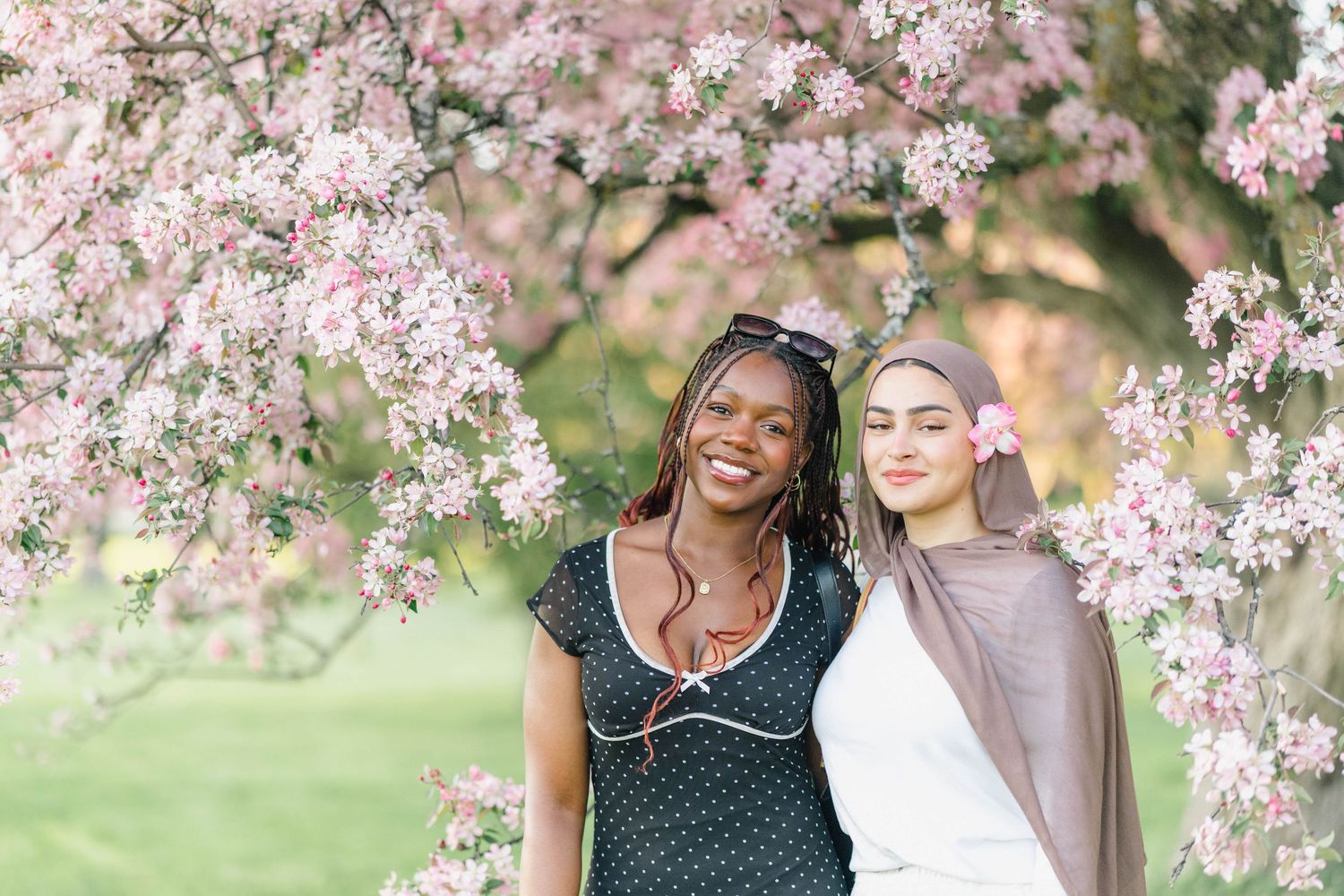 Two women smiling under pink cherry blossom tree in spring garden, one wearing hijab and floral dress.