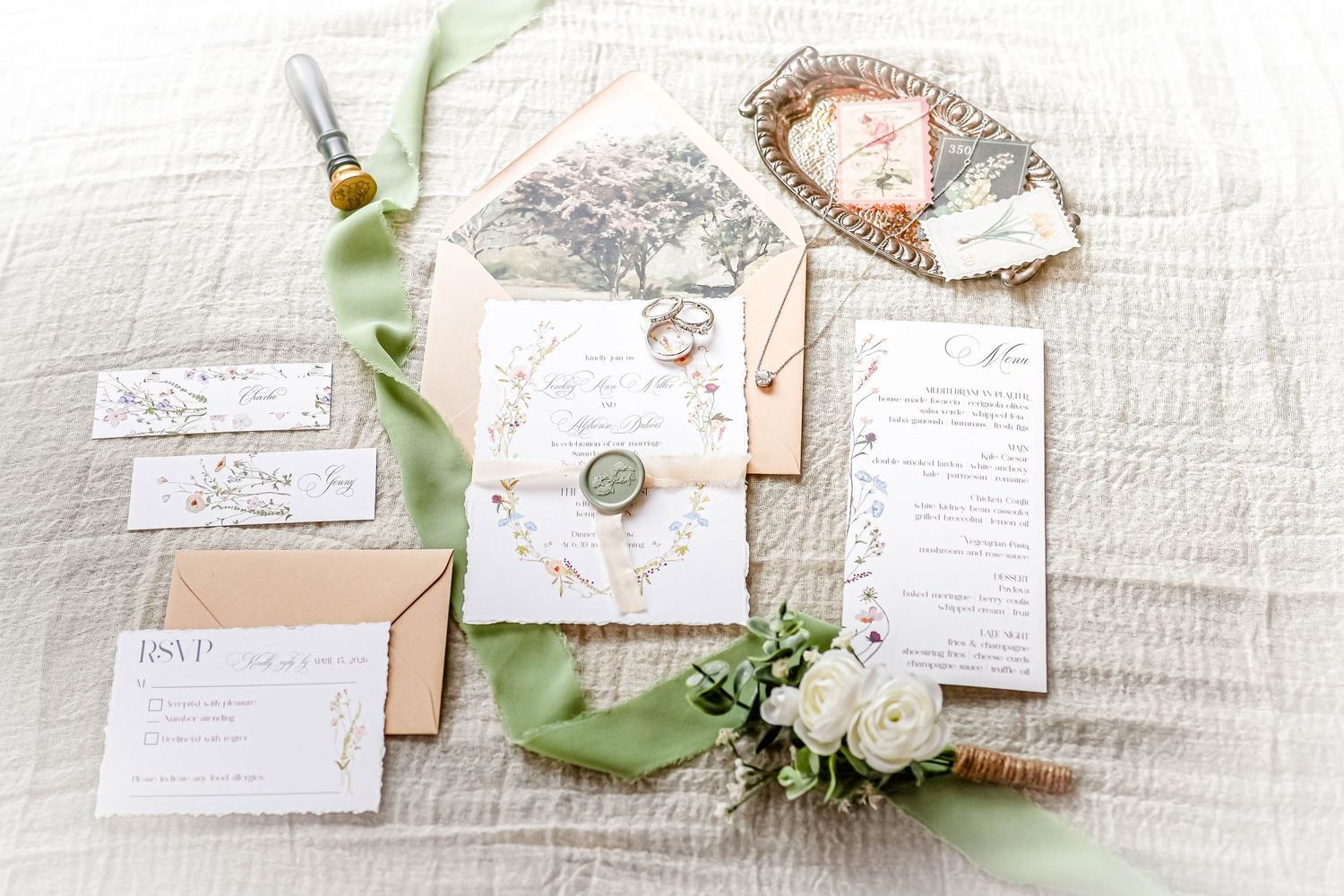 Elegant wedding invitations and stationery with floral designs and green silk ribbons displayed on white textured fabric.