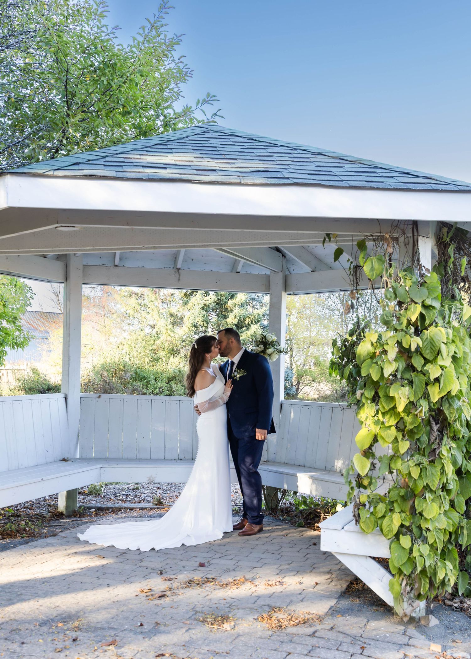 Newlyweds share intimate moments under a white pergola with climbing vines.