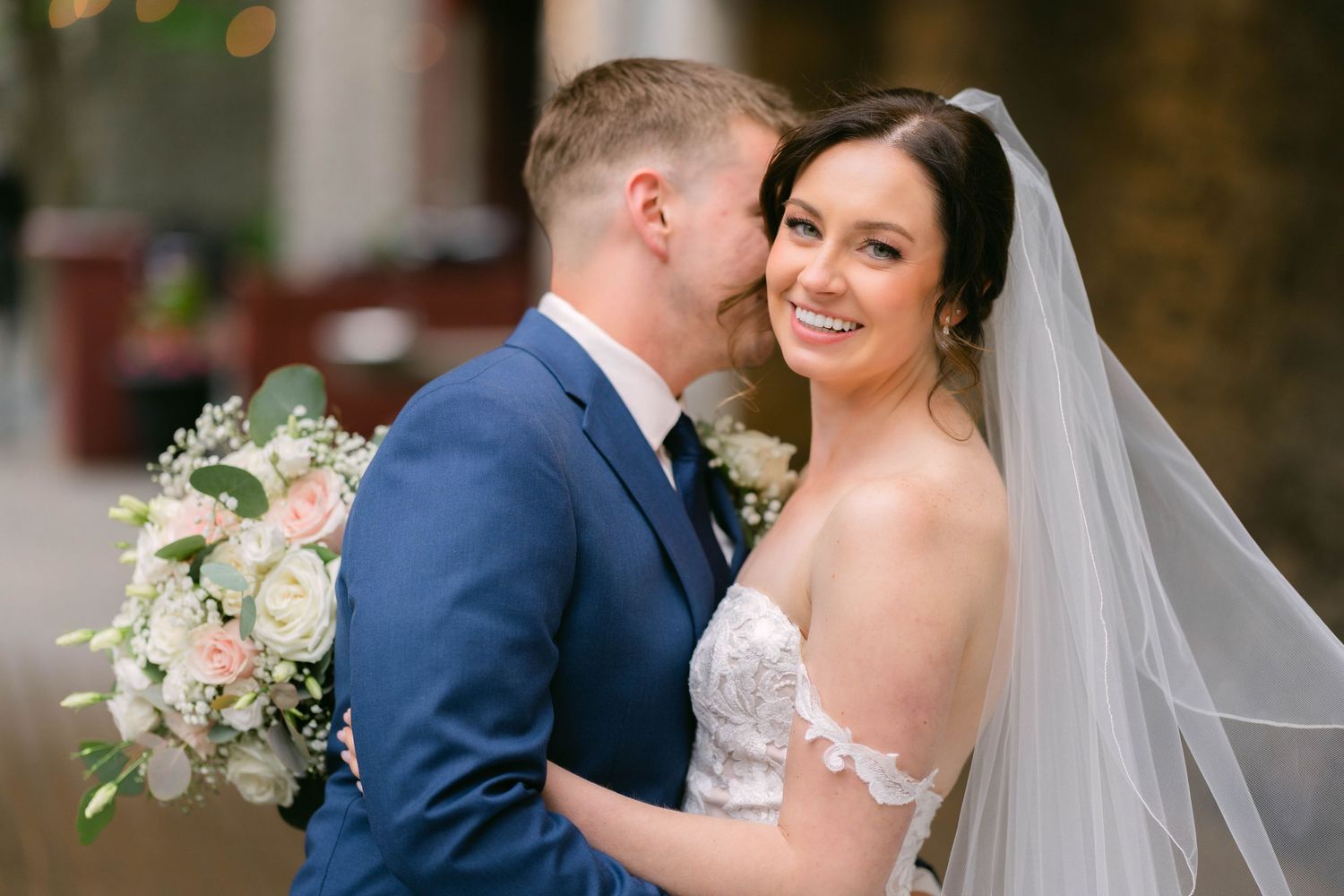 A groom in a navy suit kisses his bride's cheek as she smiles, holding a white floral bouquet on their wedding day.