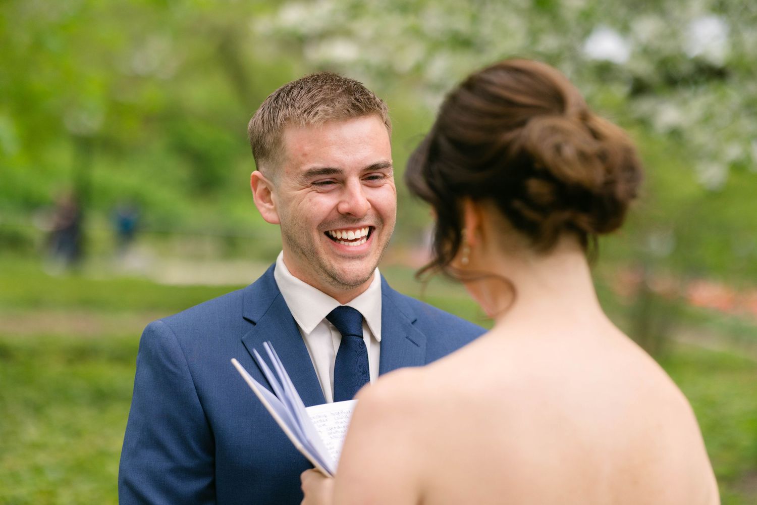 Groom in navy suit smiling joyfully at bride during outdoor wedding ceremony with lush green trees in background.