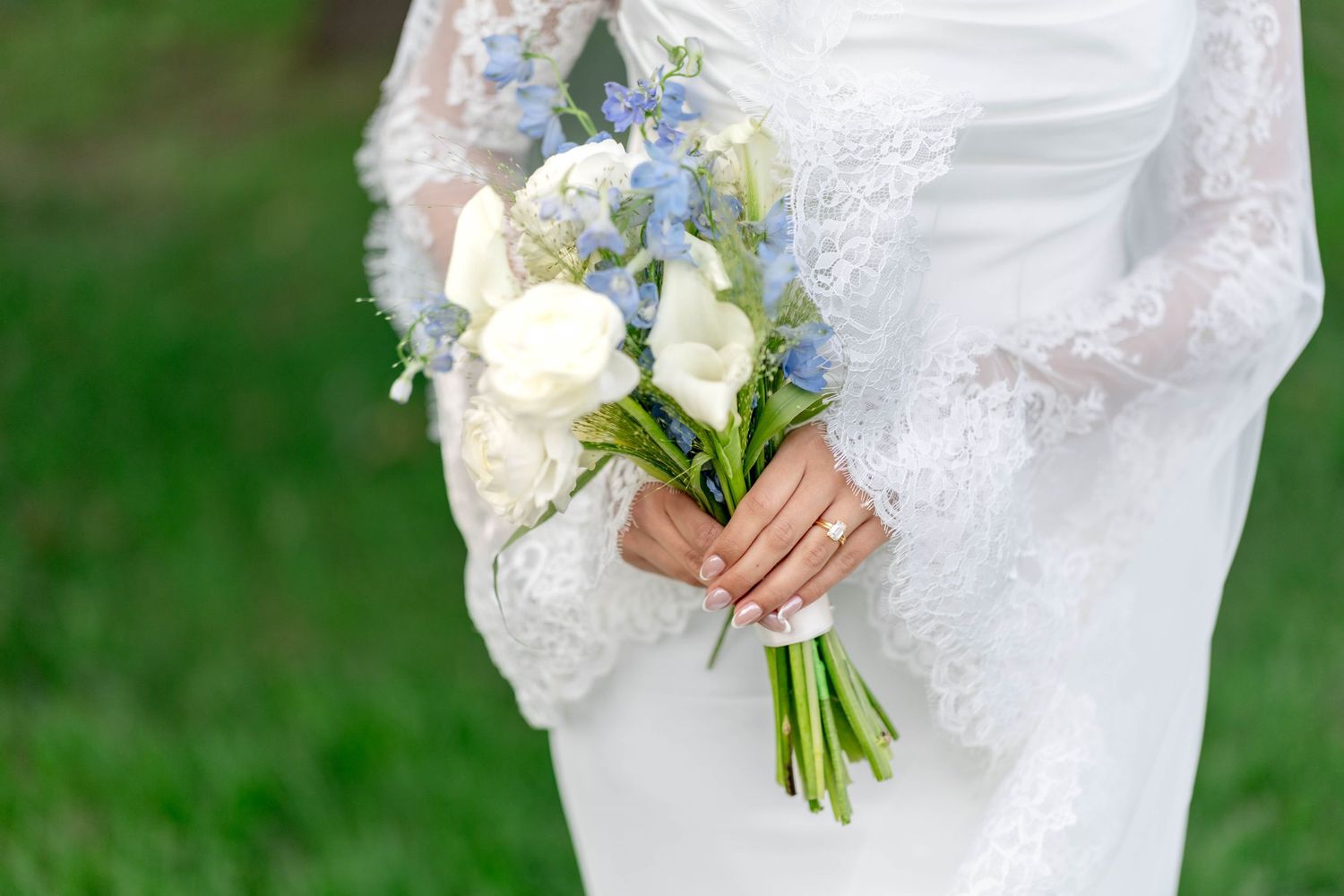 Bride in lace wedding dress holding elegant bouquet of white and blue flowers with green stems outdoors.