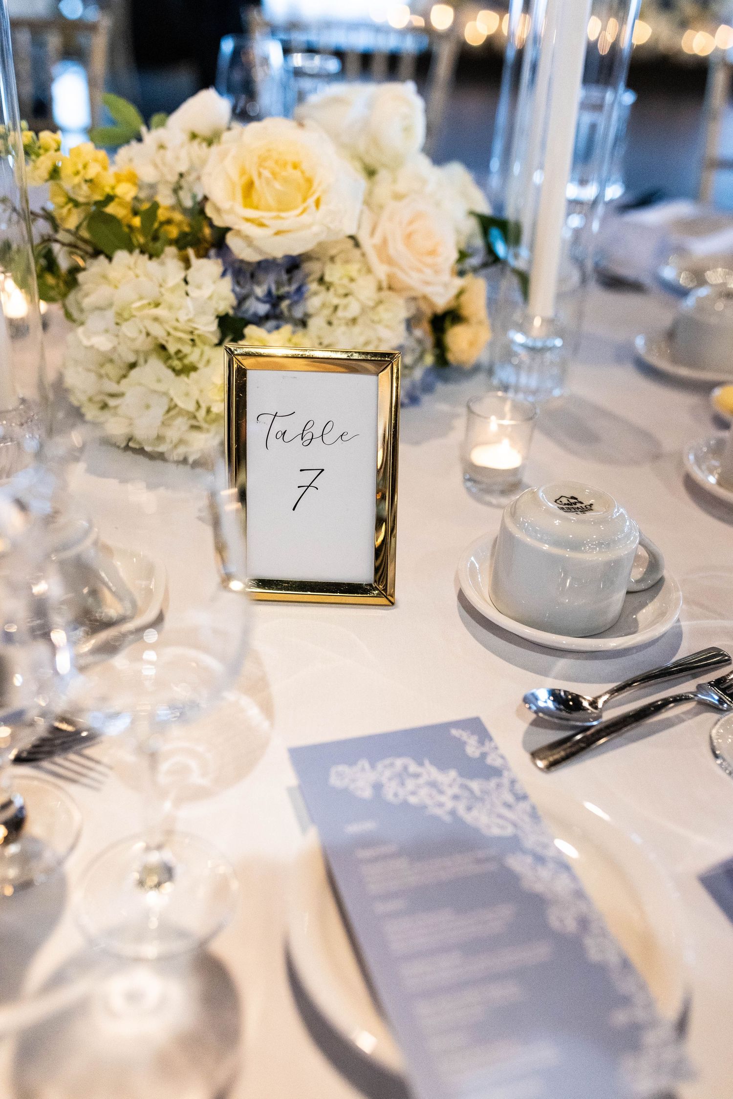 Elegant wedding table setting with gold-framed Table 7 sign, white floral centerpiece, and blue menu cards.