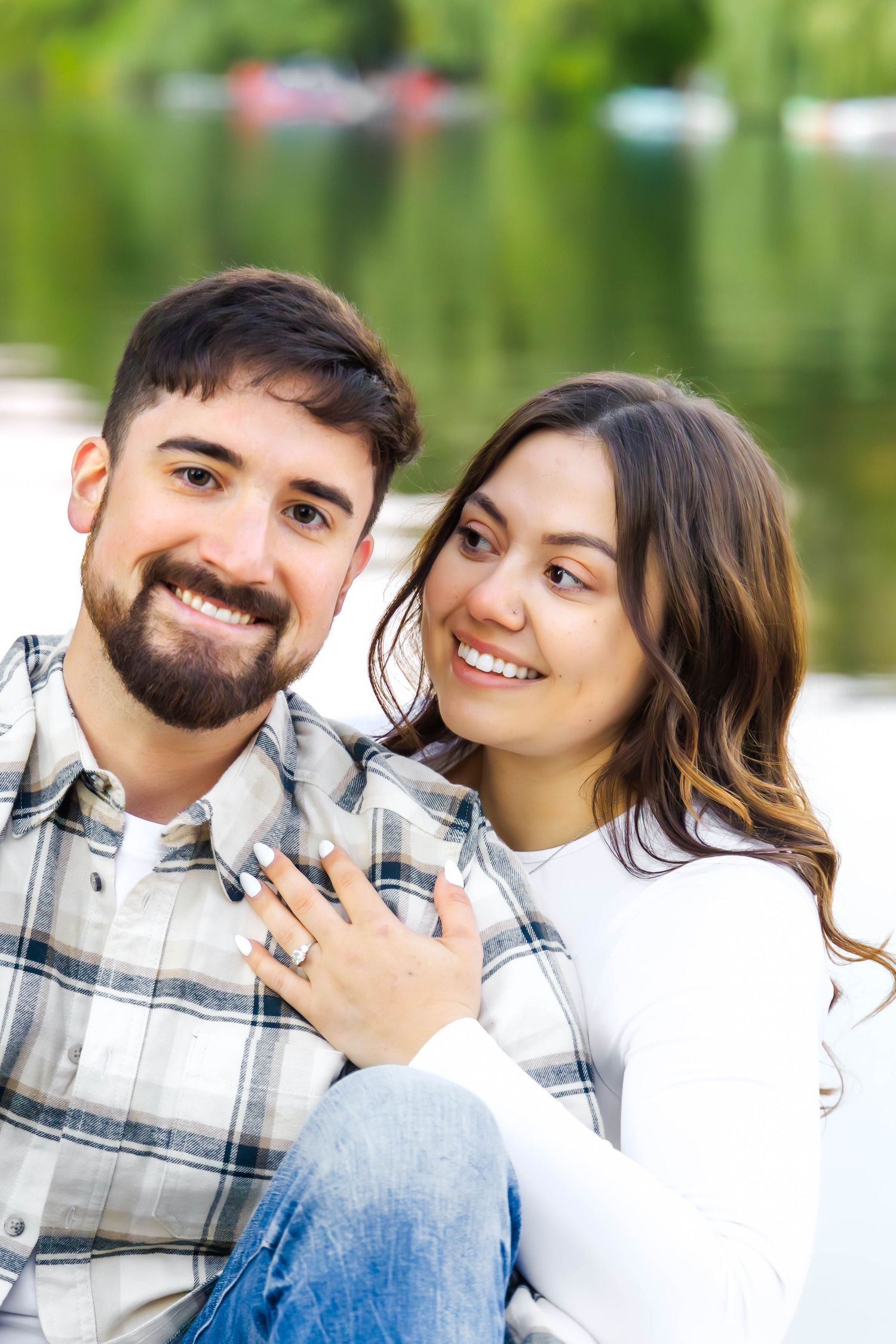 Couple in plaid and white share joyful moment by serene lake with boats in distance.