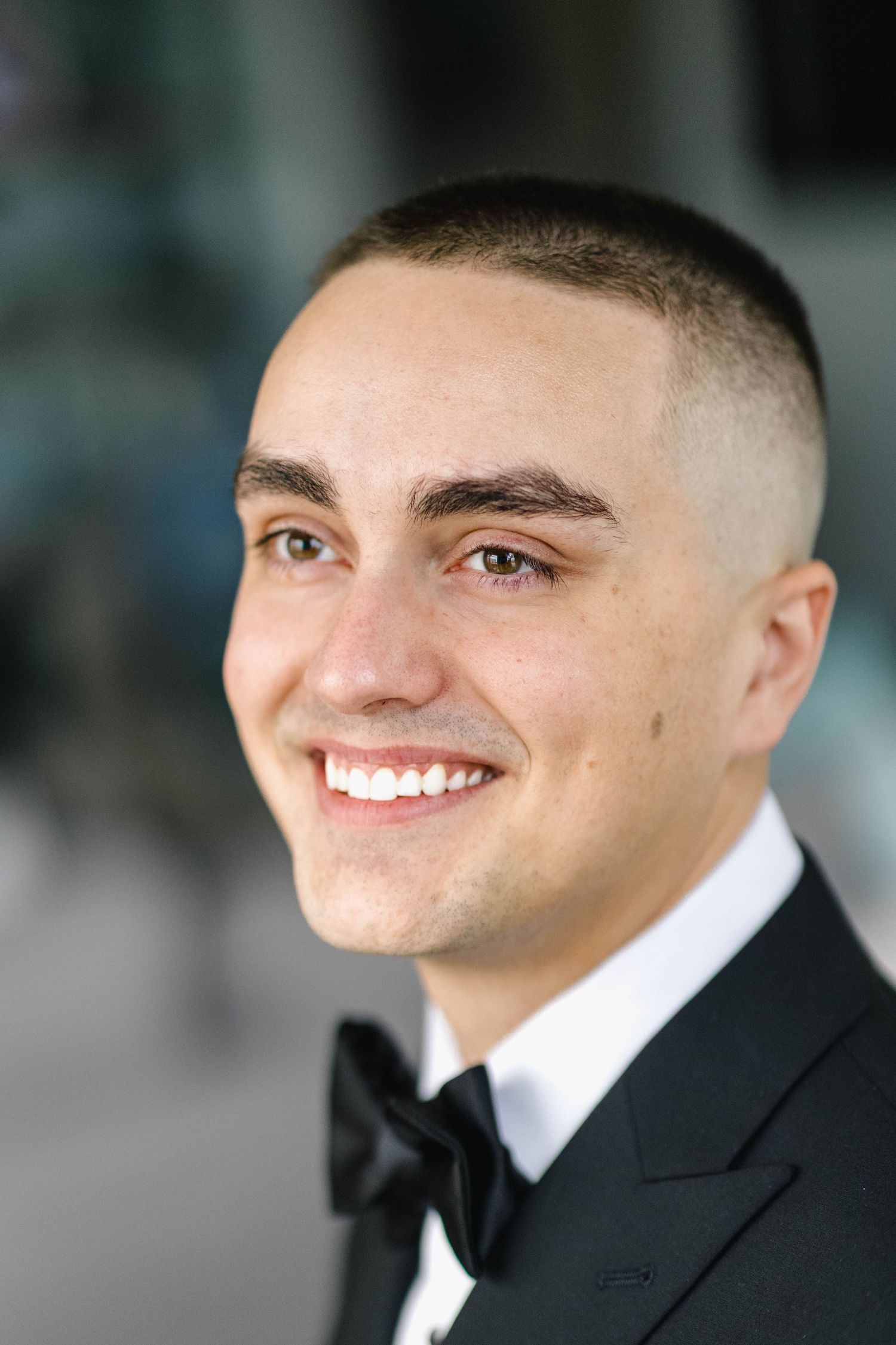 Young man in black tuxedo and bow tie smiling confidently with short buzzed hair at formal event.