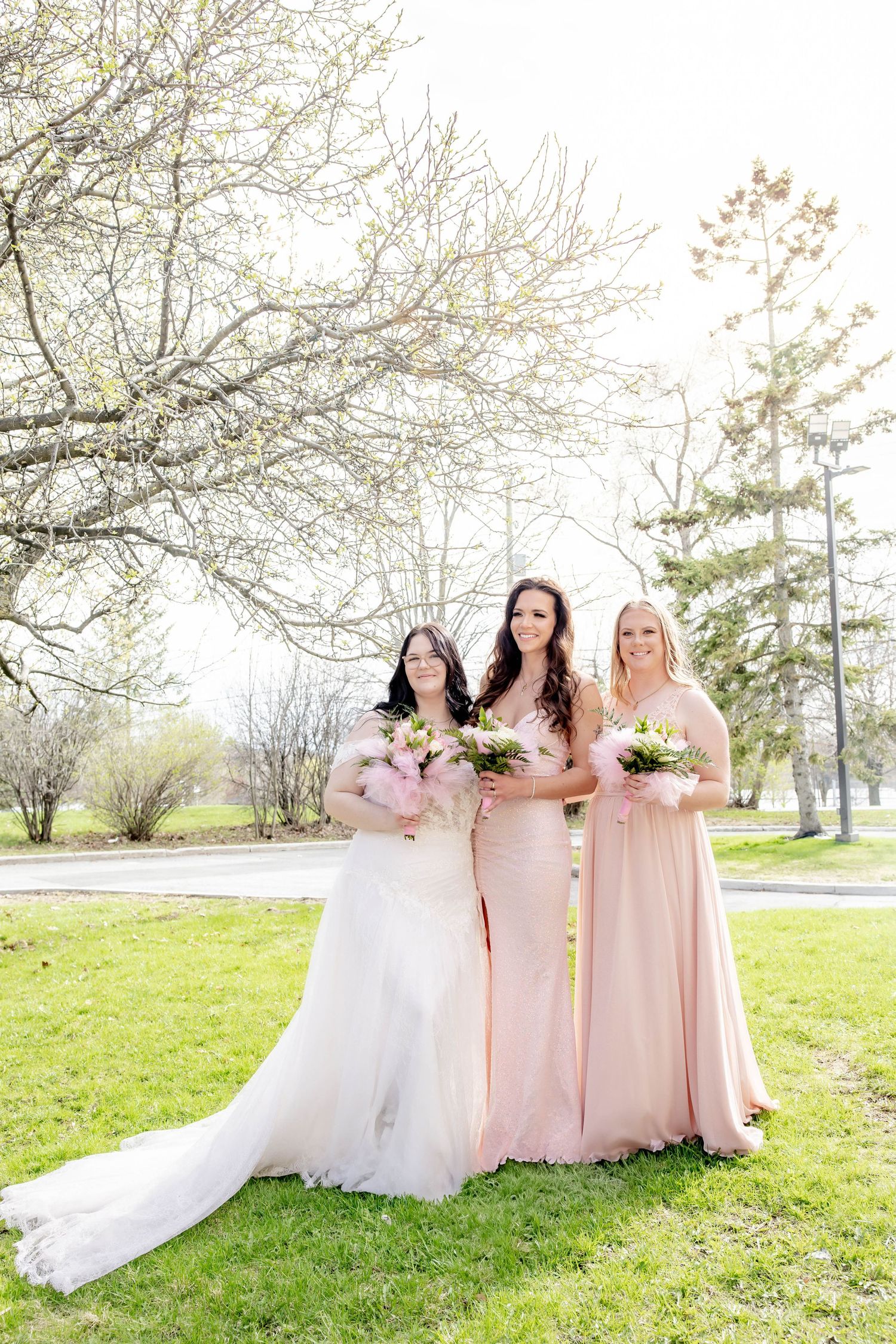 Bridal party in pink and white dresses stands together on grass under spring blossoms.