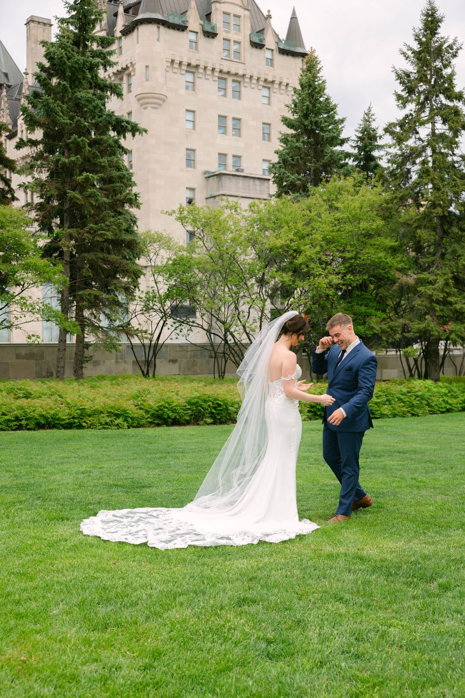 Bride and groom share a tender first look moment on lush green lawn with a grand castle-like building backdrop.