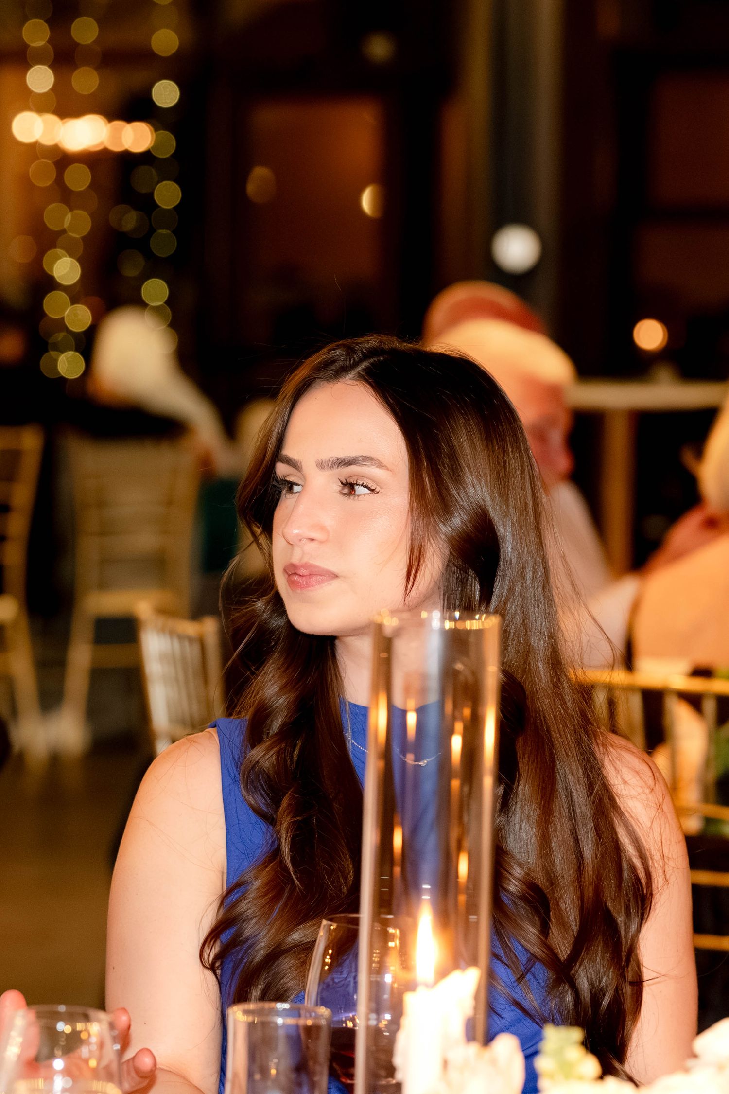 A young woman with long dark wavy hair in a blue dress sits at an elegant candlelit event table.