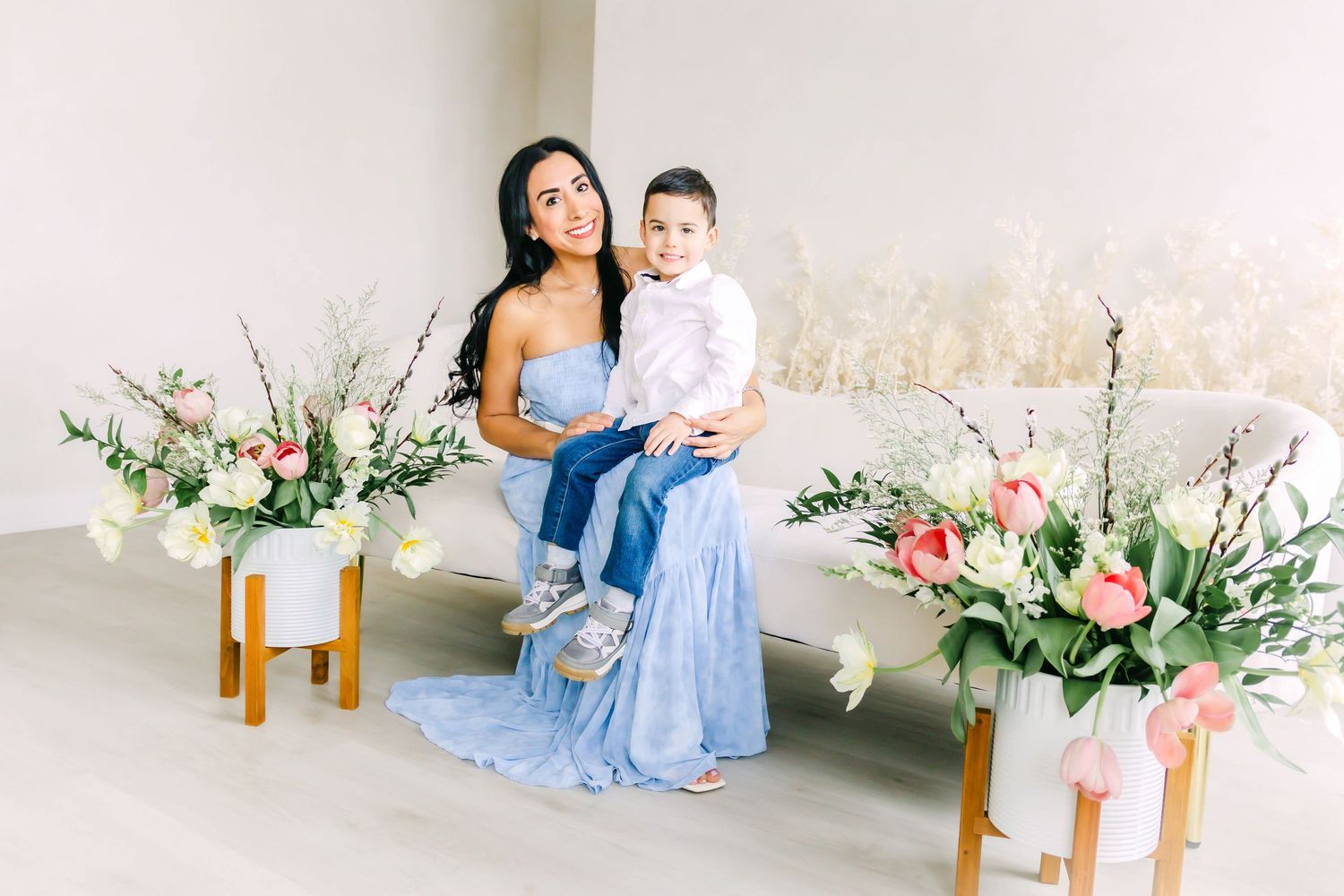 A woman in a blue dress sits on a white bench with a child, surrounded by floral arrangements on gold stands.