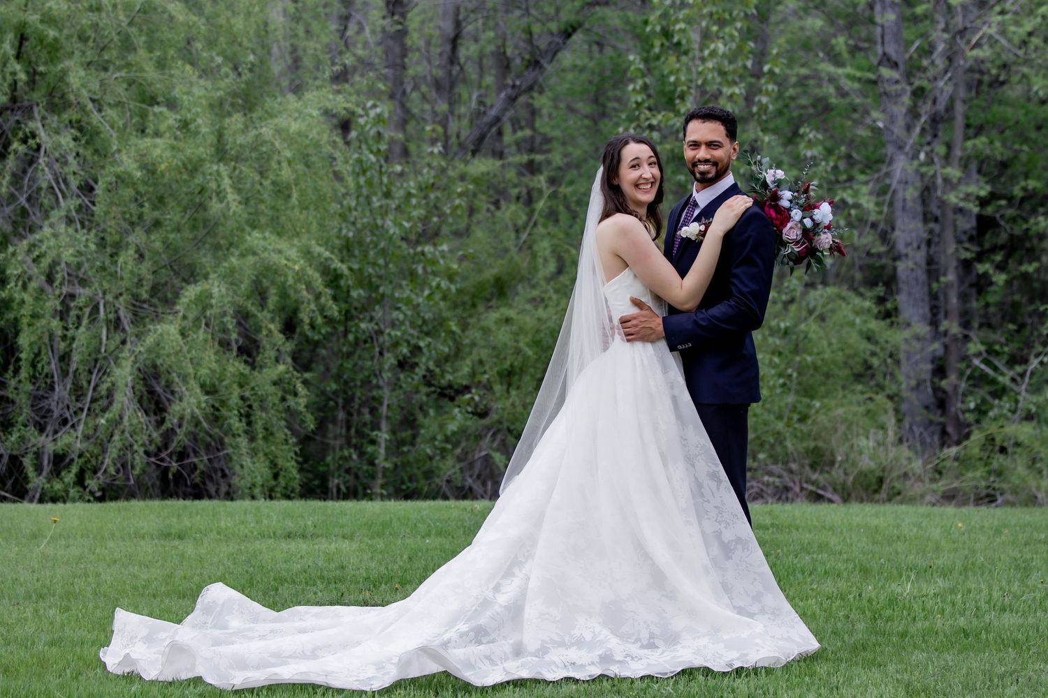 Wedding couple posing together in a lush green outdoor setting with flowing white dress.