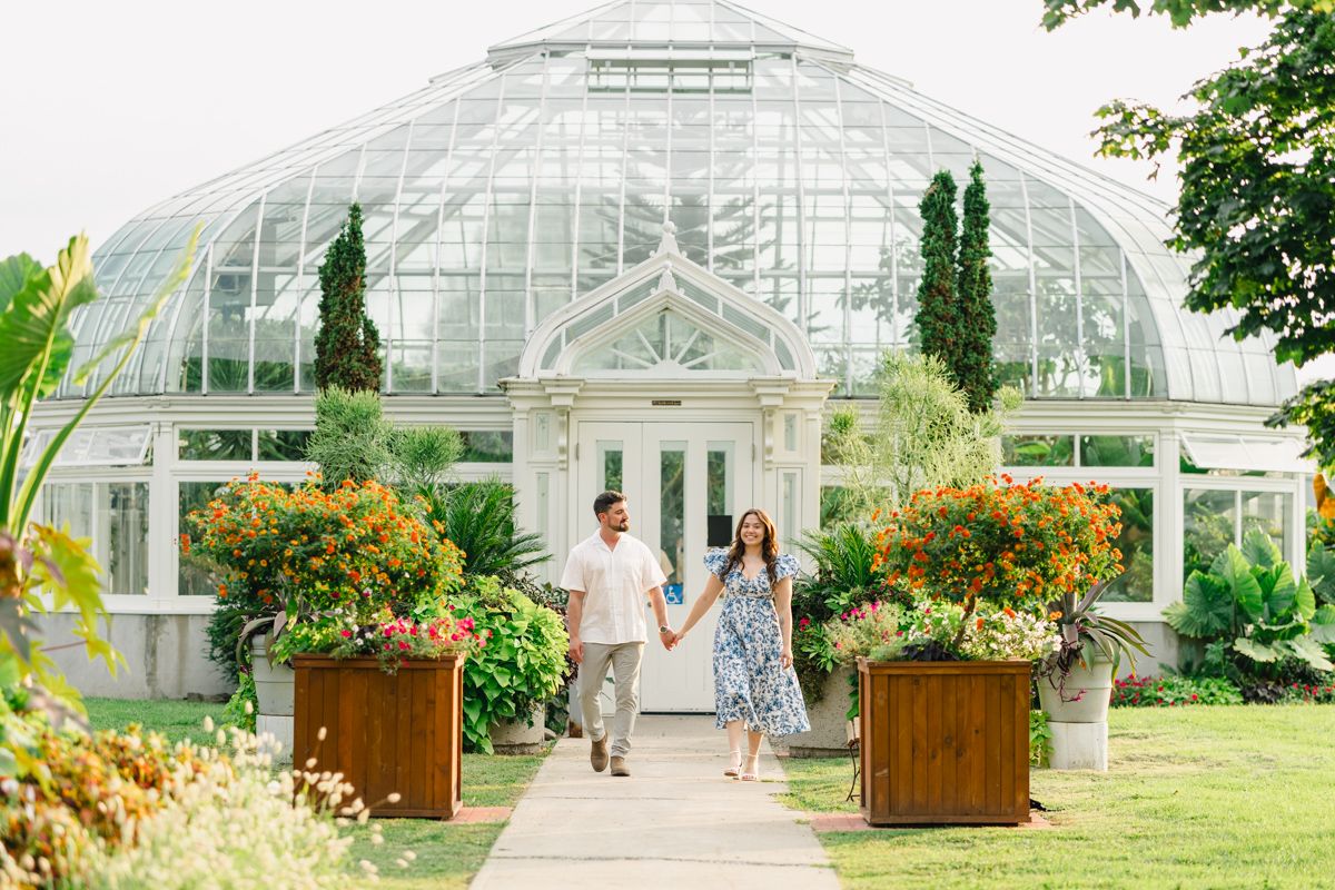 Couple holding hands walking in front of a beautiful glass greenhouse surrounded by colorful flowers.