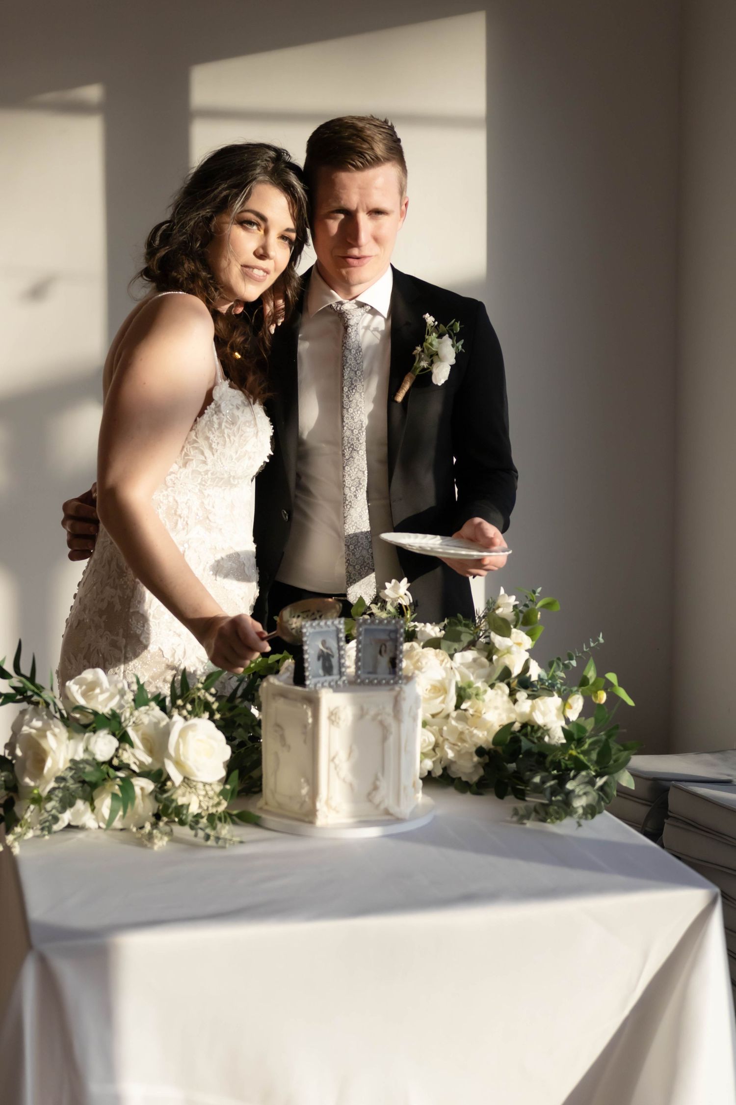 Wedding cake cutting scene with white floral decorations and dramatic window lighting.