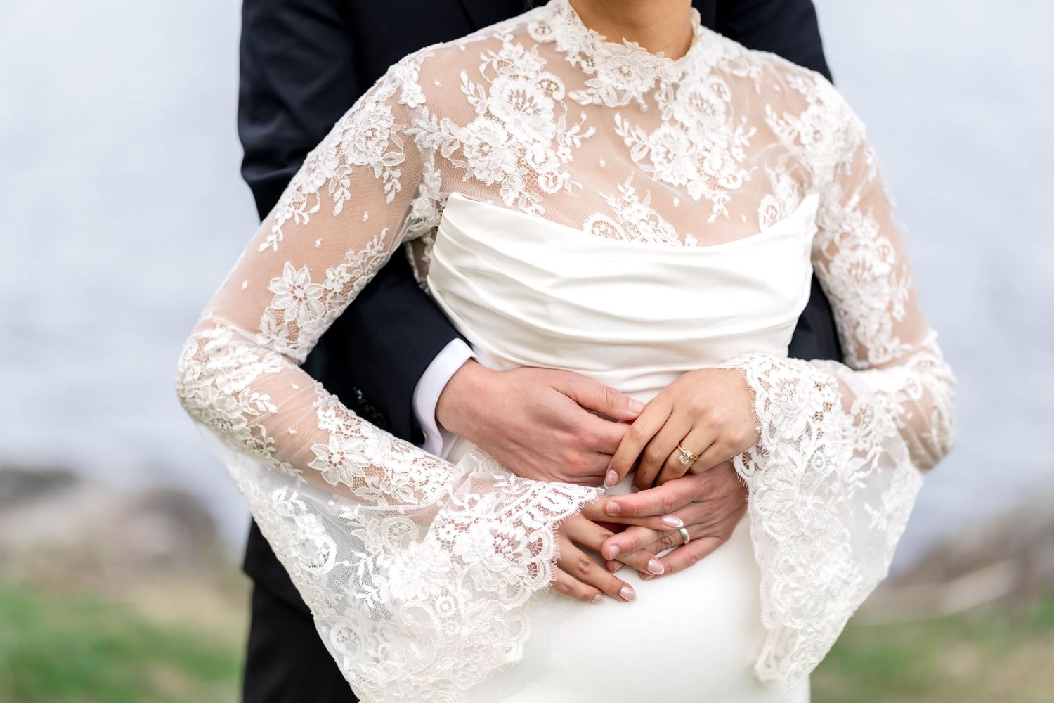 Bride in lace long-sleeve wedding gown embraced by groom in black suit, close-up of hands at waist.