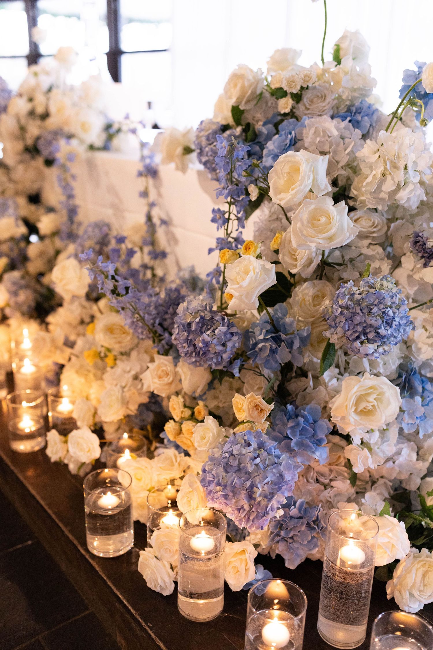 Elegant wedding table with white and blue hydrangea floral arrangements surrounded by glowing candles in glass holders.