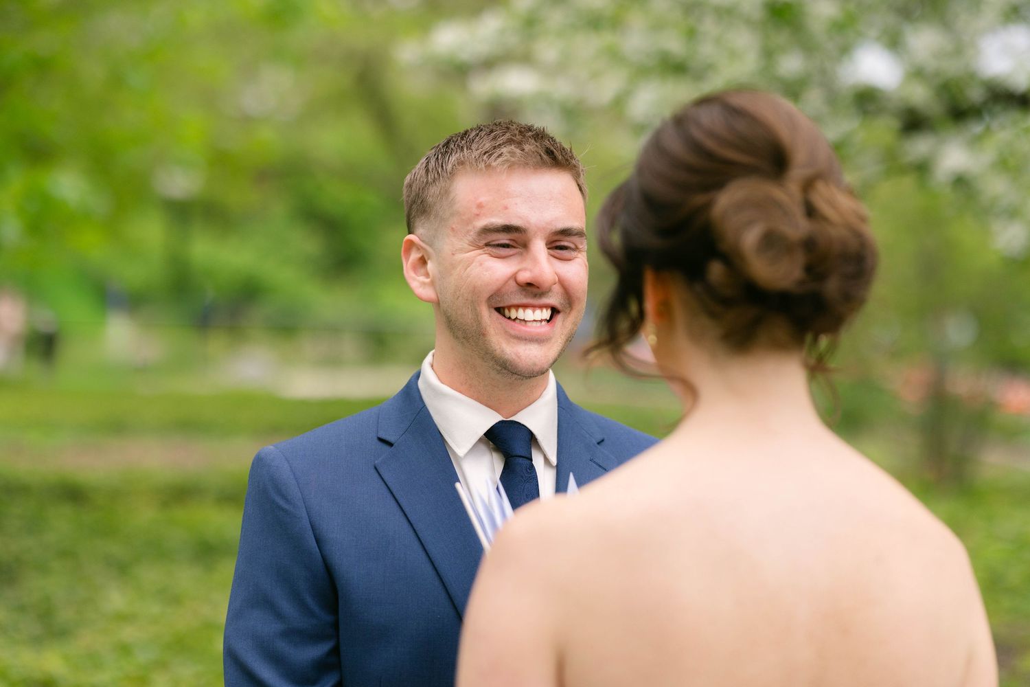 Groom in navy suit smiling at bride during outdoor wedding first look ceremony surrounded by lush green trees.