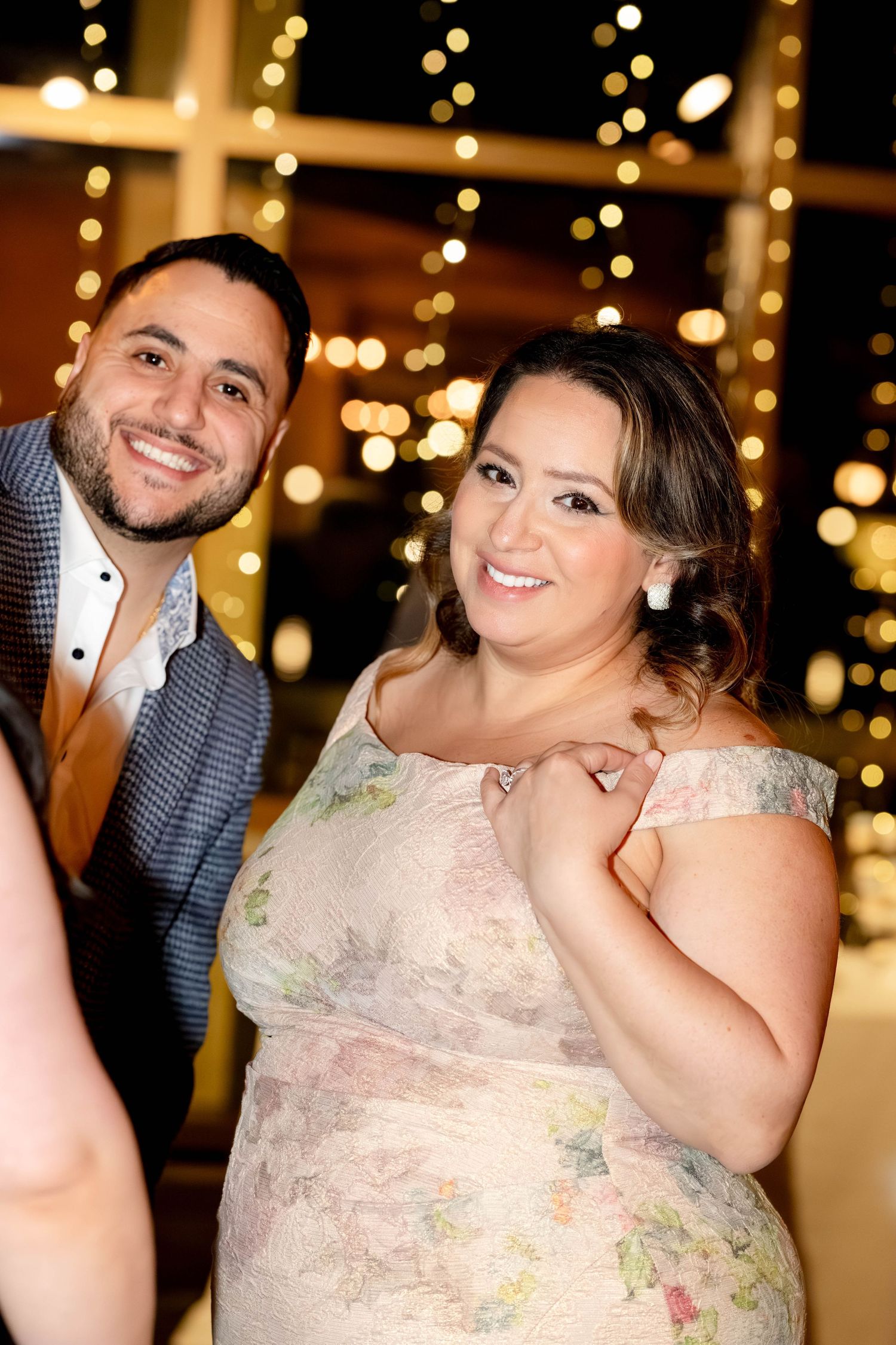 A smiling couple poses together at an elegant evening event with warm fairy lights glowing in the background.